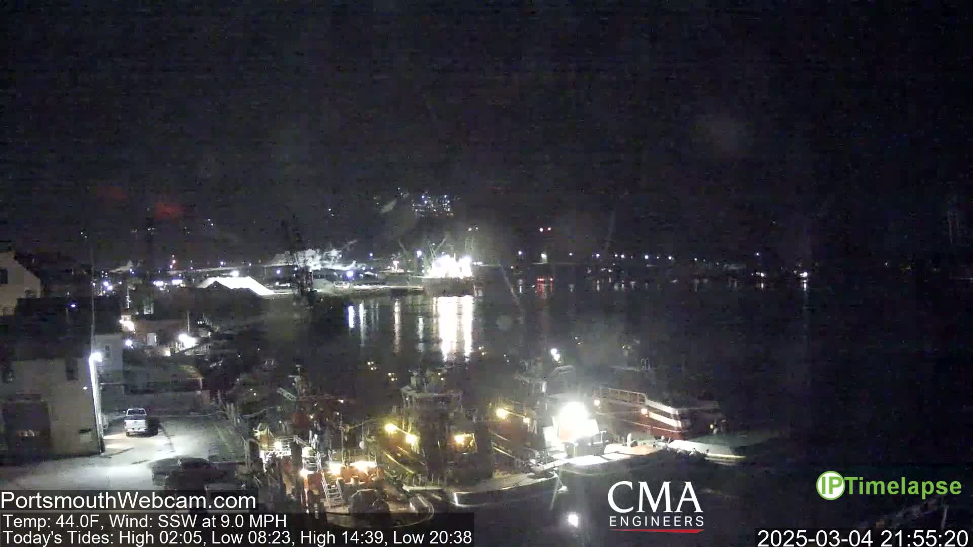 A nighttime harbor scene shows numerous boats docked along a waterfront, under a dark, clear sky with a gentle south-southwesterly wind.