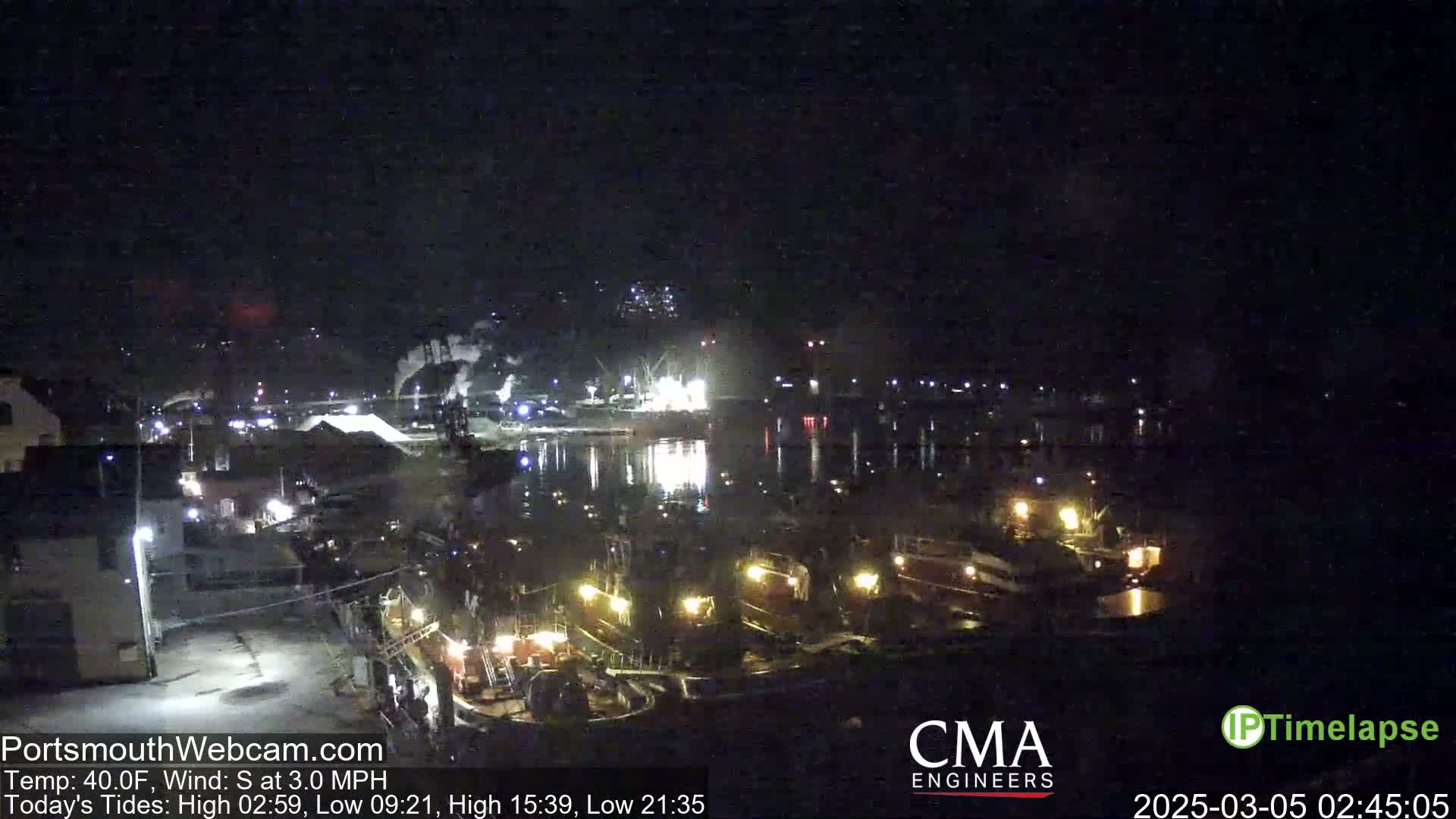 A nighttime view of a harbor with several docked boats illuminated, under a dark sky with a south wind.