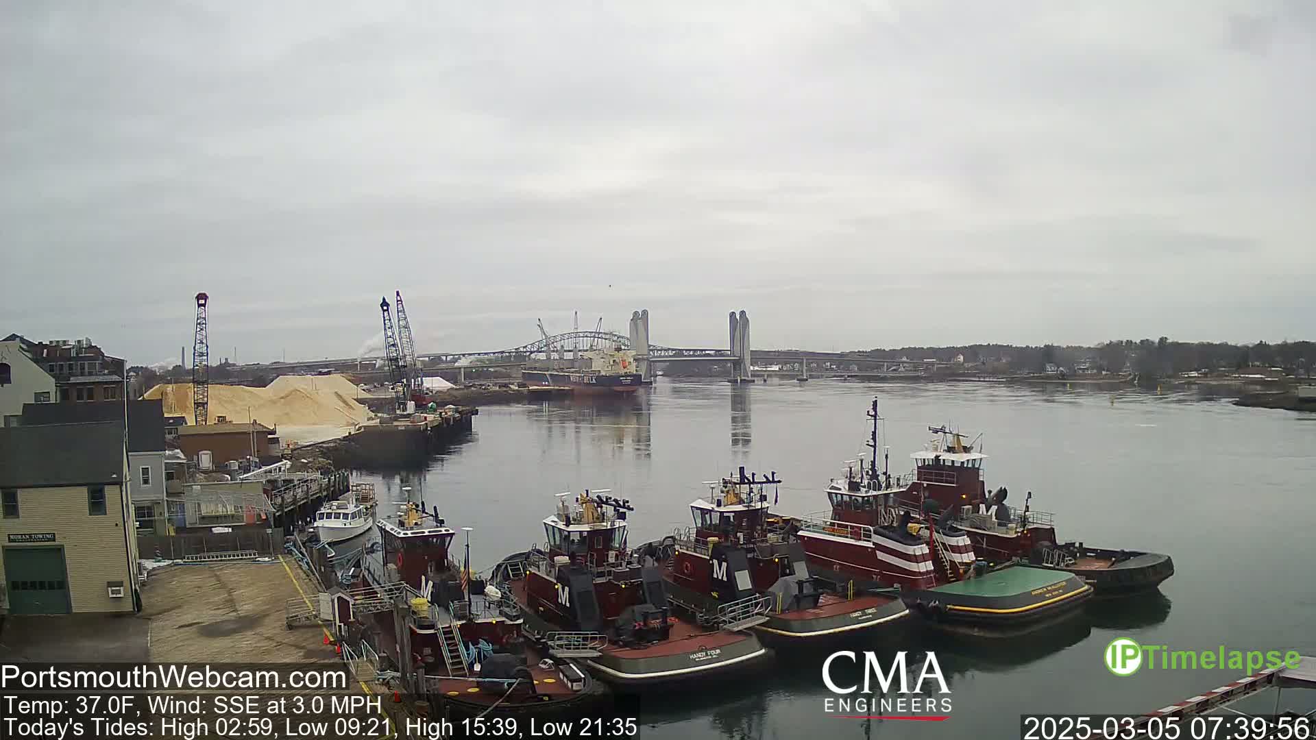 Several tugboats are moored at a dock next to a large pile of sand, with a bridge and a larger vessel visible in the distance on a cloudy day.