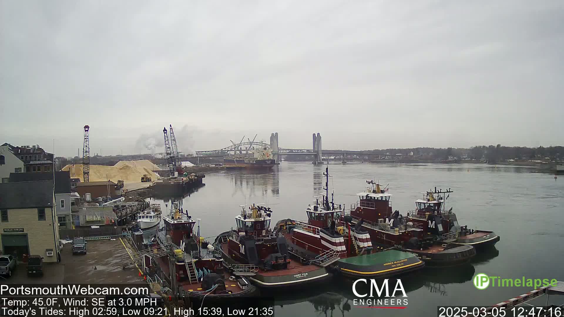 Several tugboats are moored at a dock next to a large pile of sand, with a bridge and a cargo ship visible in the distance on an overcast day.