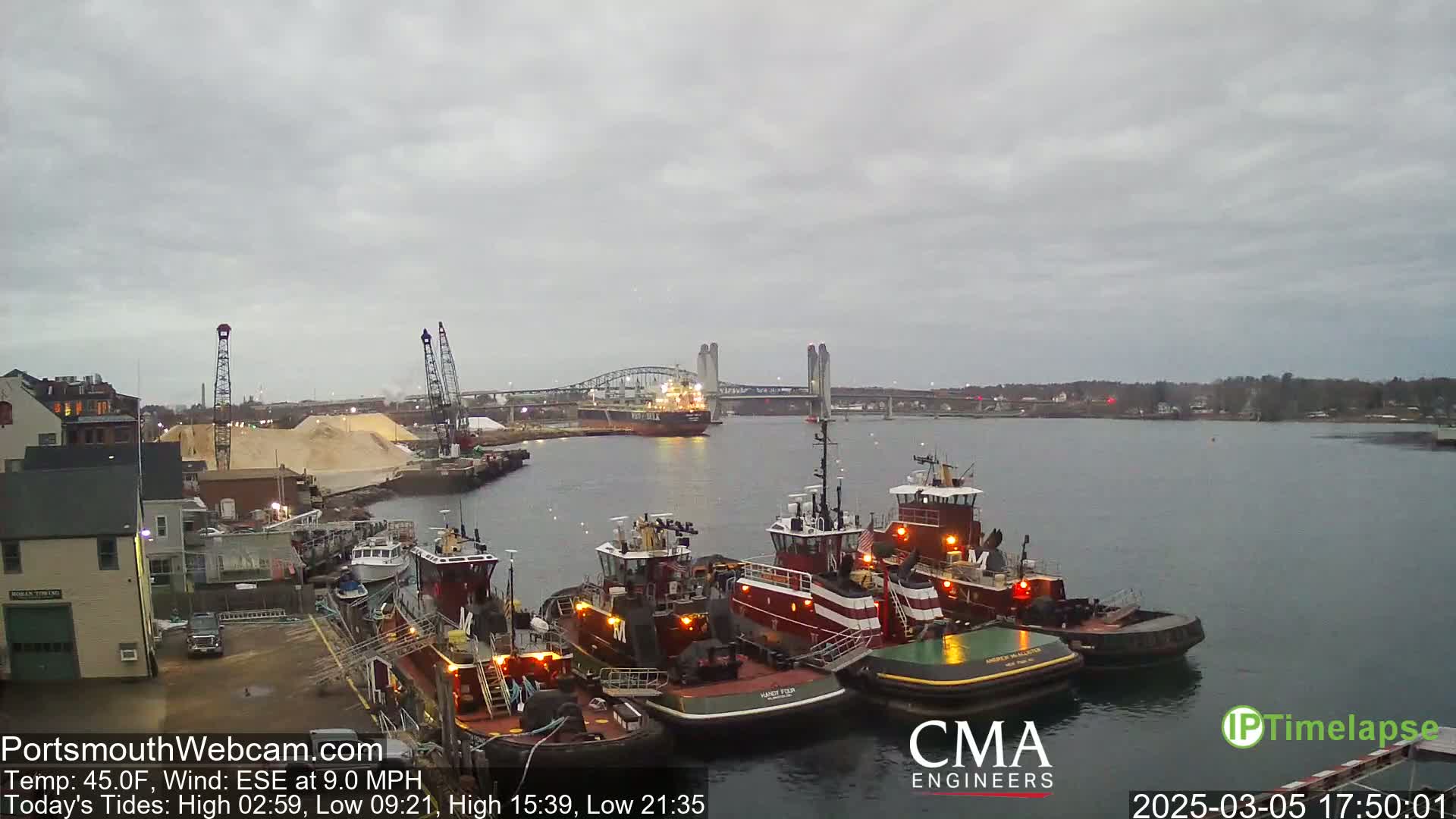 Several tugboats are docked at a waterfront, under an overcast sky, with a large cargo ship and a bridge visible in the distance.