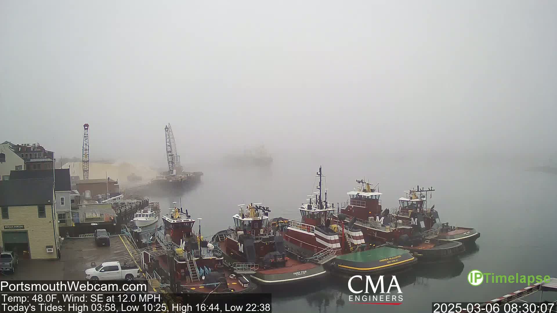 Several tugboats are moored at a dock in a thick fog, with a crane and other vessels visible in the distance.