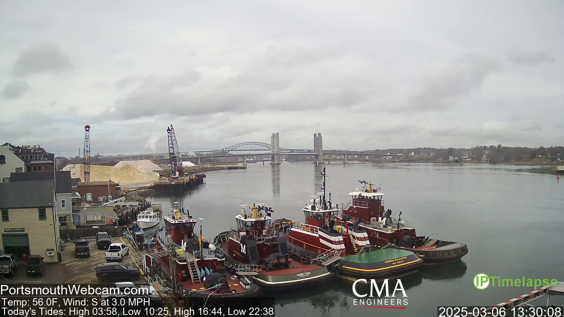Several tugboats are moored at a waterfront dock under a cloudy sky, with a bridge spanning a river in the background.