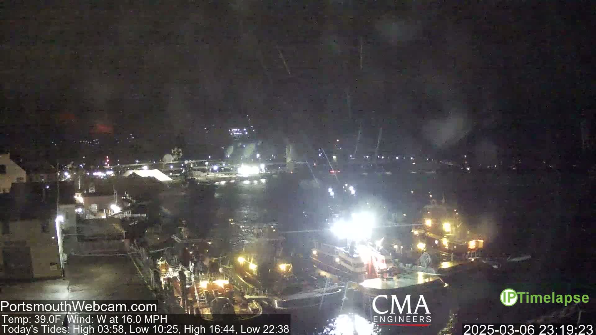 A nighttime harbor scene shows several boats illuminated by lights under a dark, slightly overcast sky with a moderate west wind.