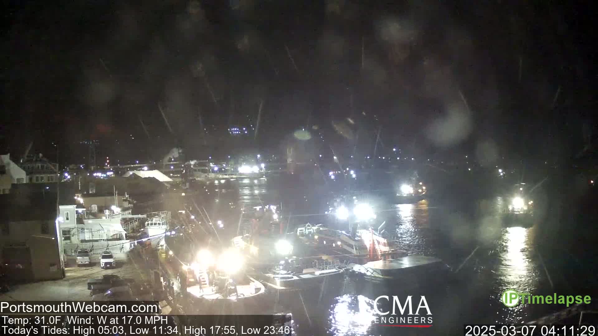 Several boats are illuminated in a harbor at night during snowy weather.