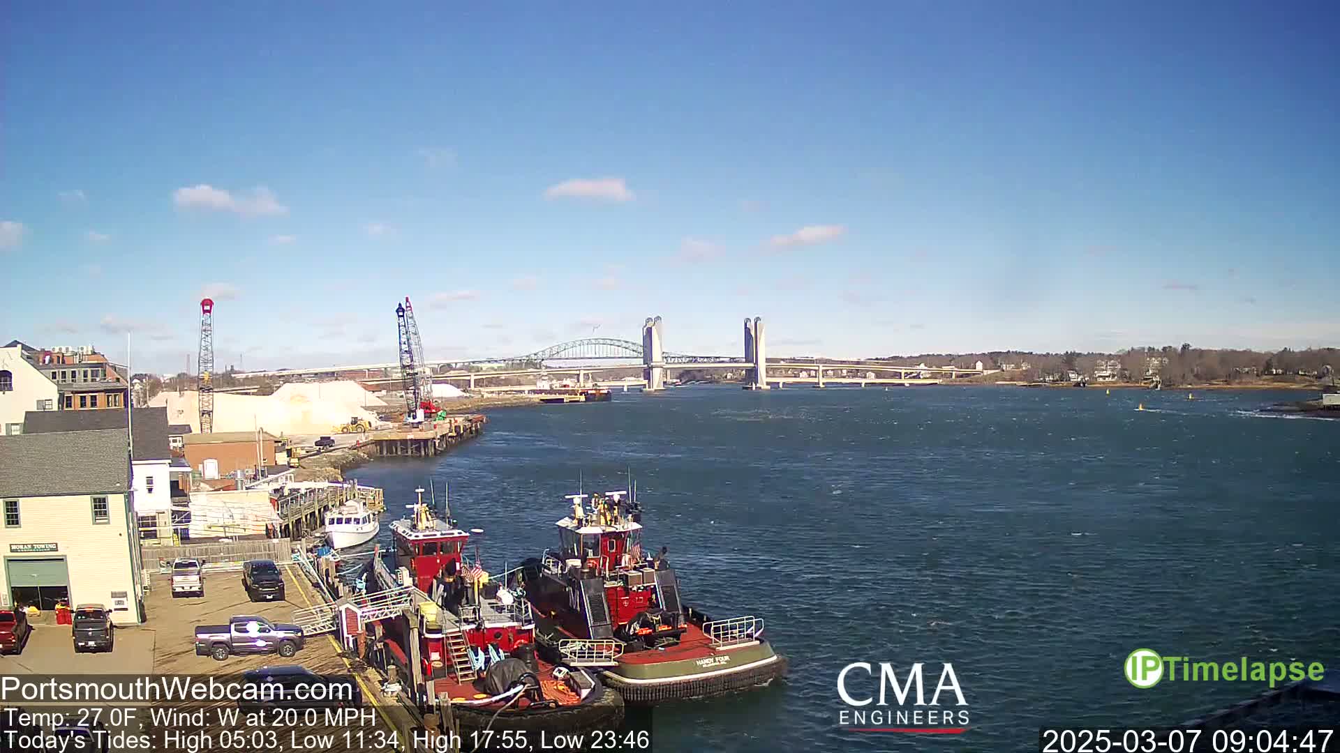 Two tugboats are docked near a waterfront with a bridge and a large pile of sand in the background, under a partly cloudy sky with a west wind at 20 mph.