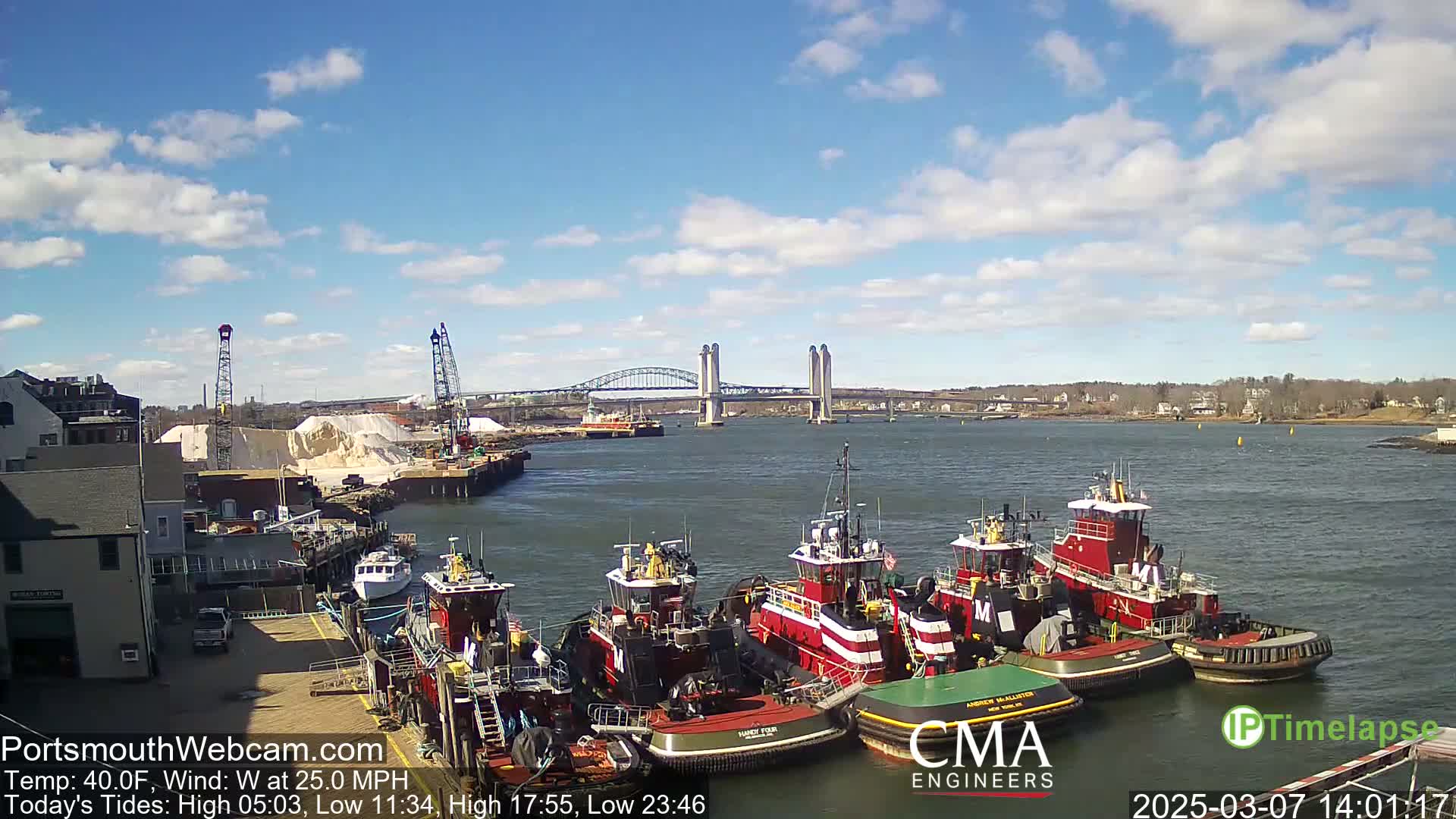 Several tugboats are docked at a waterfront, with a large bridge and a pile of what appears to be sand or salt in the background under a partly cloudy sky.