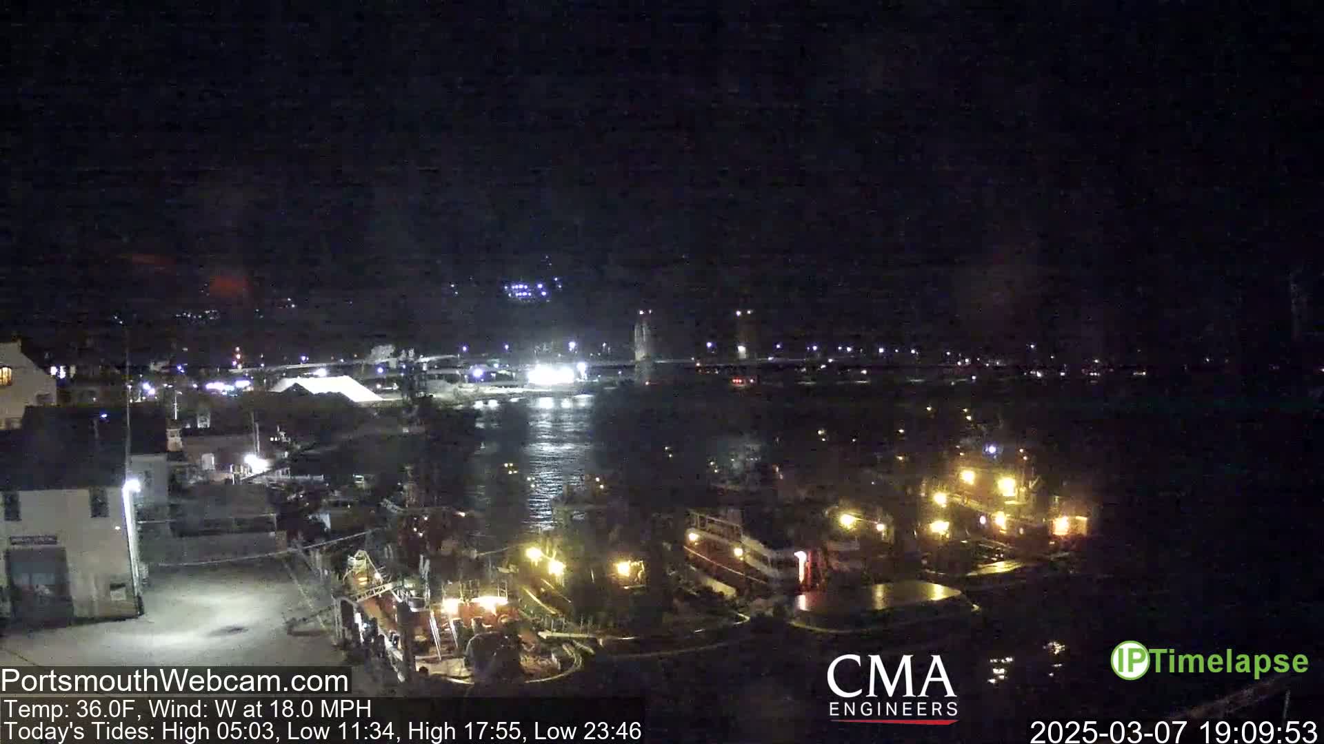 A nighttime harbor scene shows several docked boats illuminated by lights, a bridge in the distance, and calm water under a dark sky.