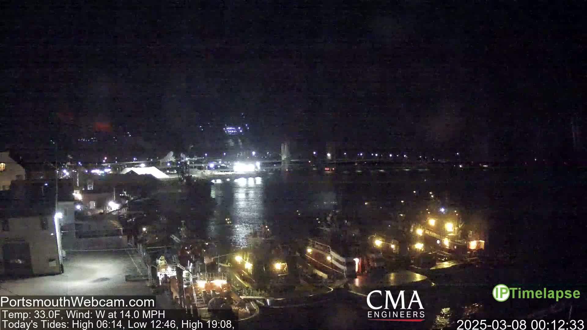 A nighttime view of a harbor with several boats docked, a bridge in the distance, and calm water under a clear, dark sky.