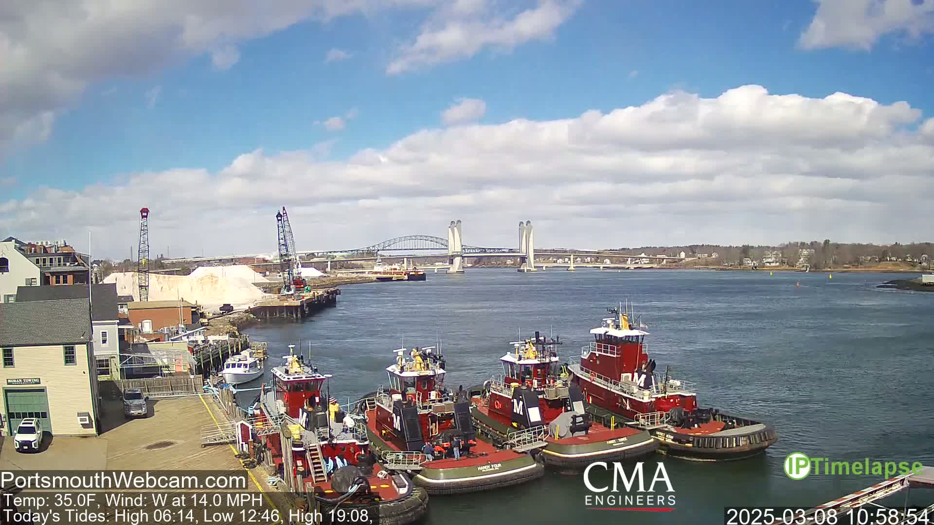 Several tugboats are moored at a dock next to a body of water, with a bridge and a partly cloudy sky in the background.