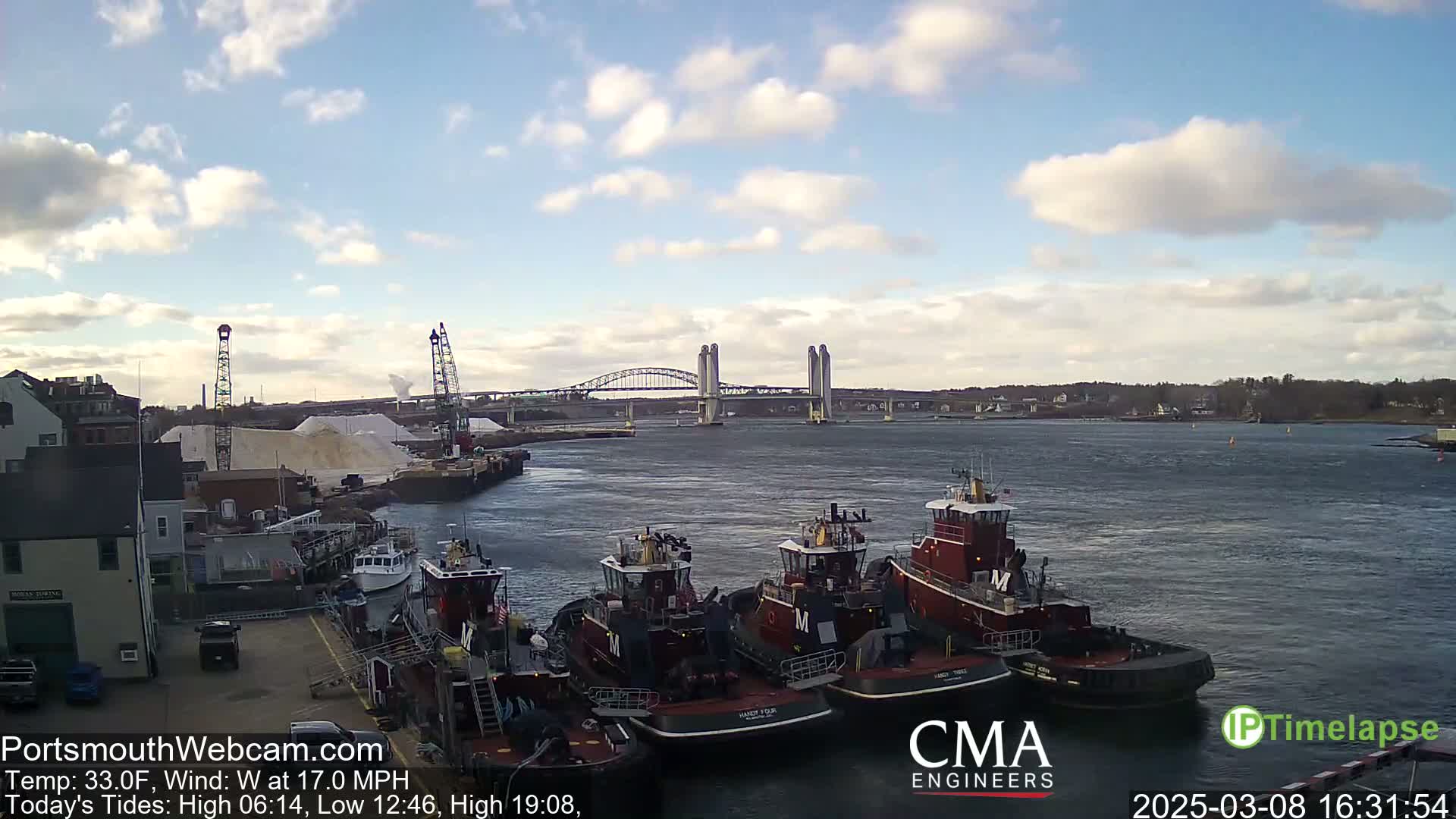 Several tugboats are docked at a waterfront, with a bridge and partly cloudy skies in the background.
