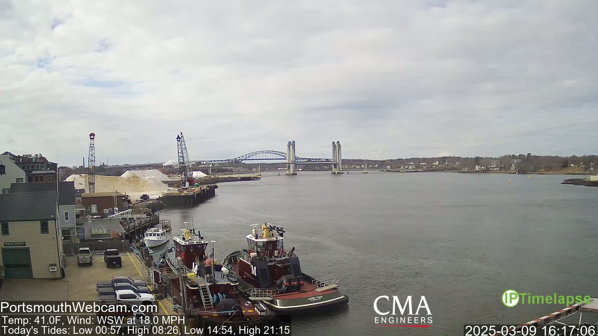 A view of a harbor with two tugboats docked near a building and a large pile of sand, a lift bridge in the background under an overcast sky.