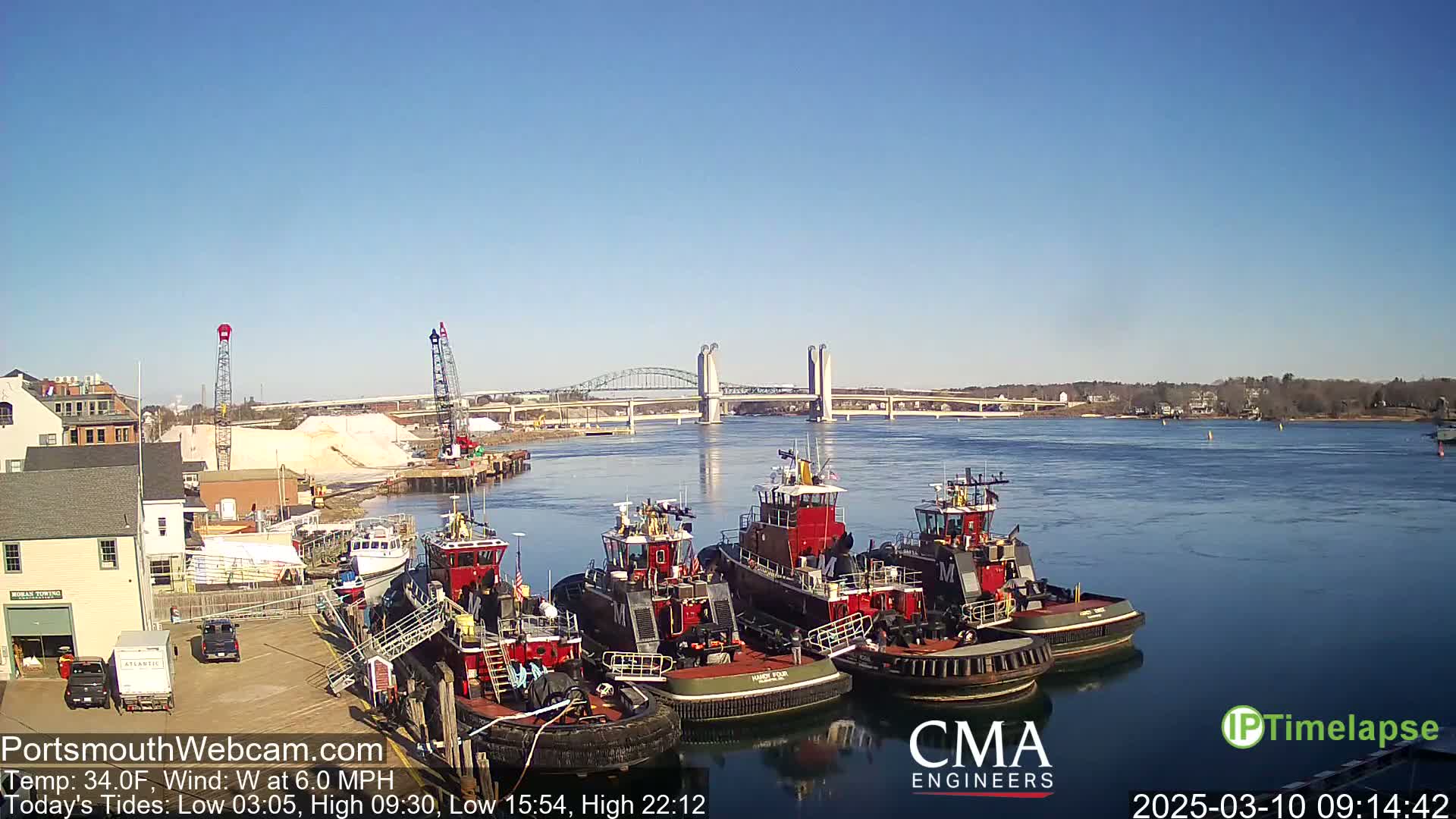 Several tugboats are docked at a waterfront location under a clear blue sky, with a lift bridge visible in the distance across a calm body of water.