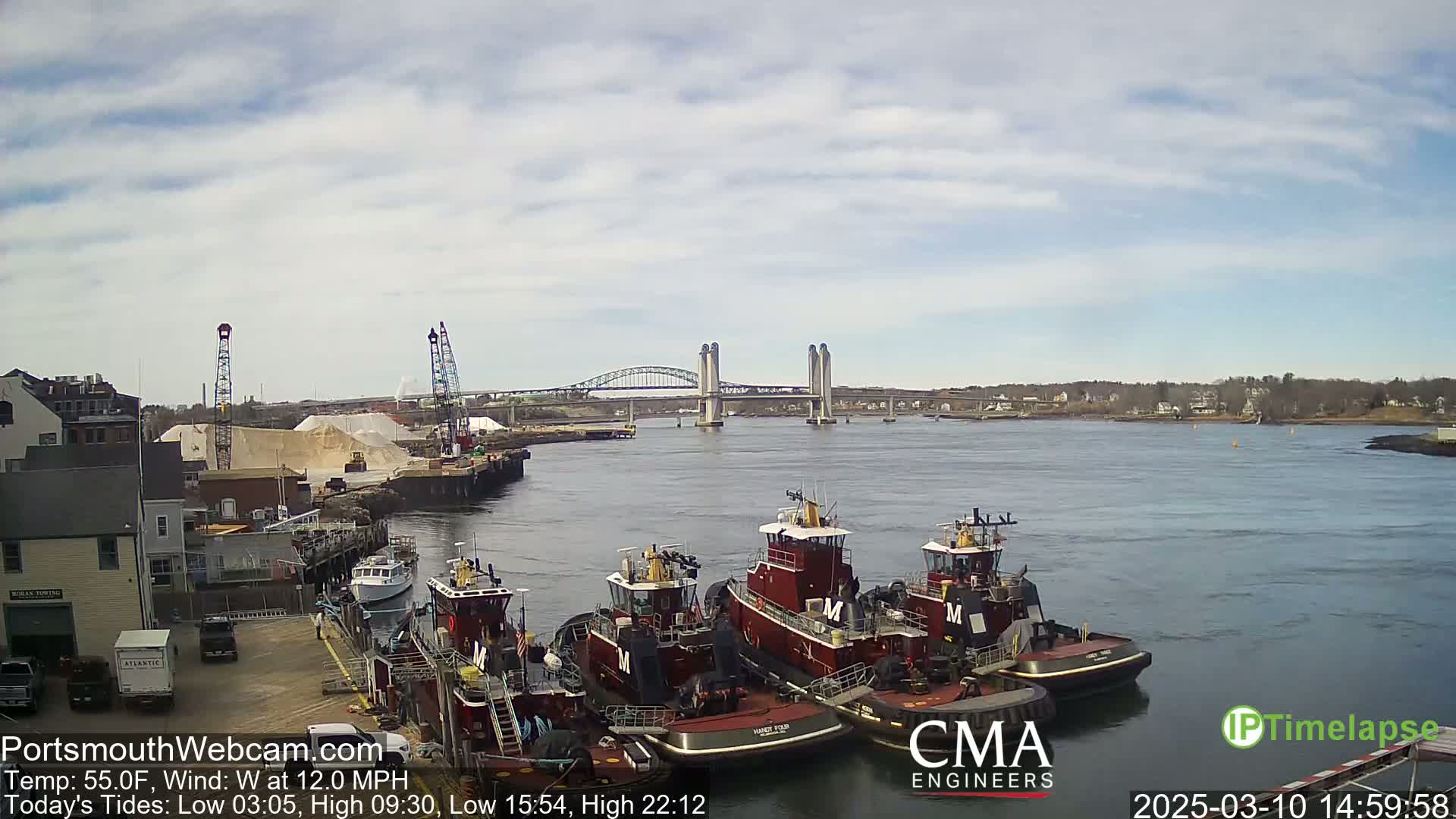 Several tugboats are docked at a waterfront industrial area, with a bridge and a large pile of sand visible in the background under a partly cloudy sky.