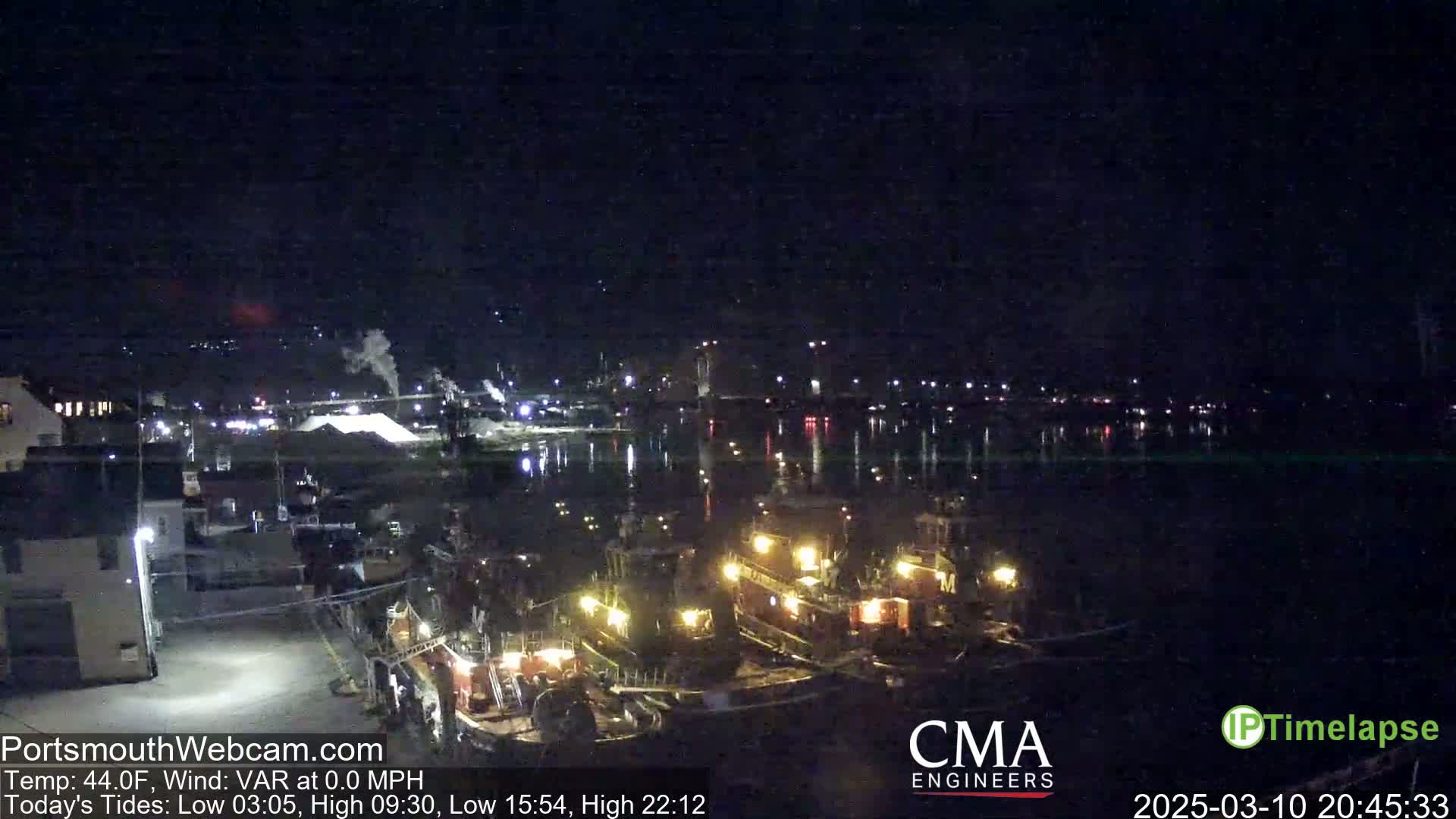 At night, several tugboats are docked near a waterfront, with calm water reflecting the city lights in the background under a clear, dark sky.