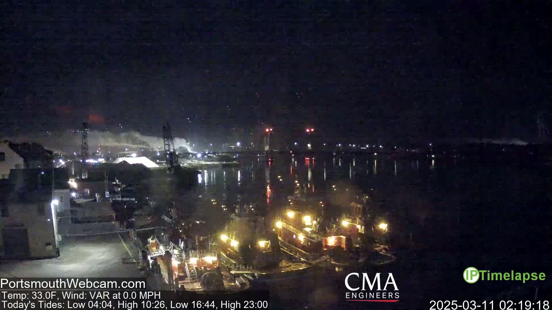 Several tugboats are docked at a nighttime harbor under a clear, dark sky, with industrial activity visible in the background.