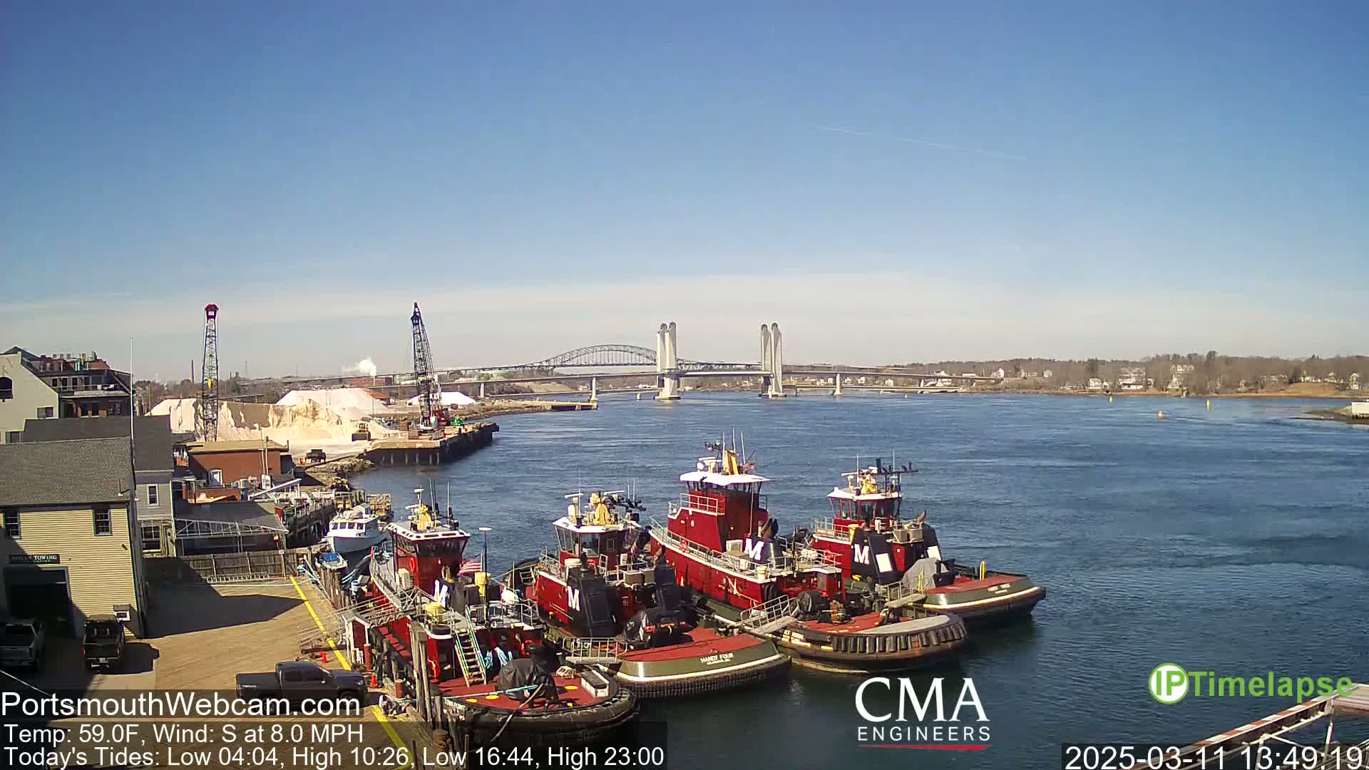 Several red tugboats are docked at a waterfront, with a lift bridge in the background under a clear, sunny sky.