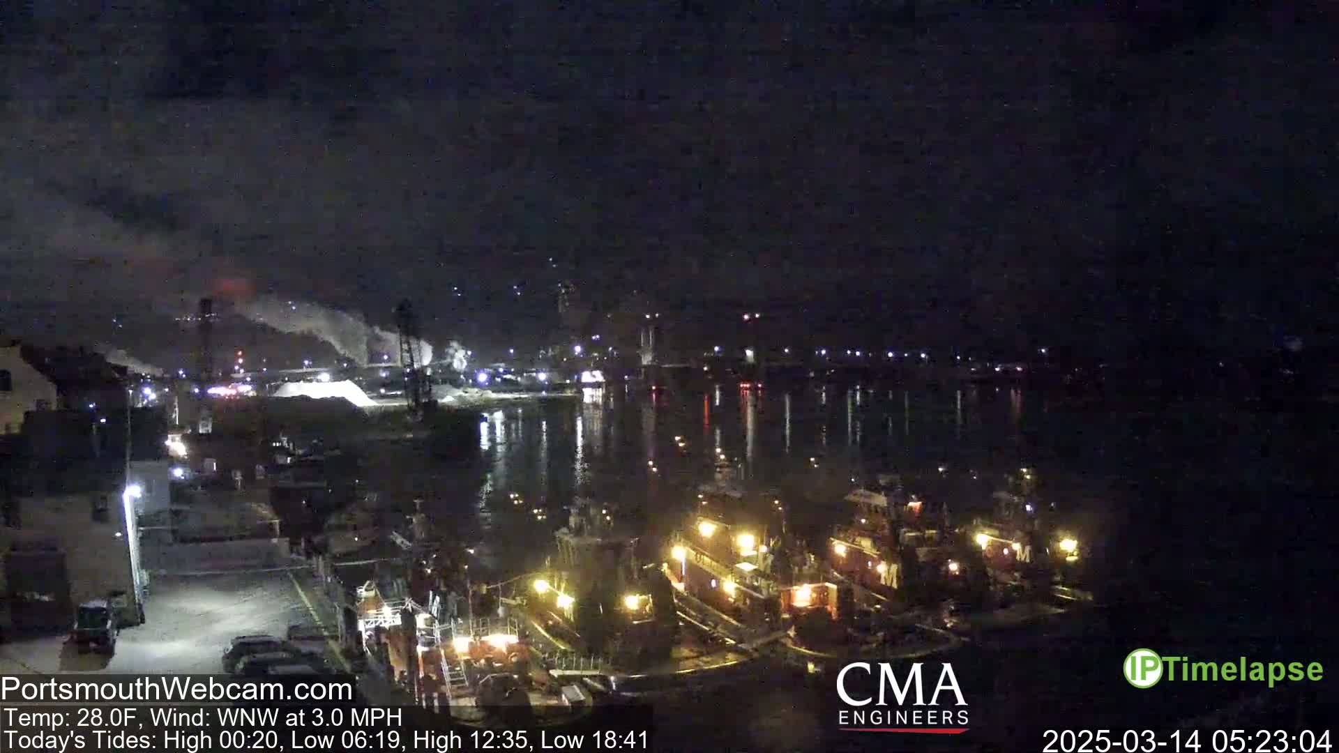 Several tugboats are moored at a nighttime dock near an industrial area with smoke stacks emitting steam under a dark sky.