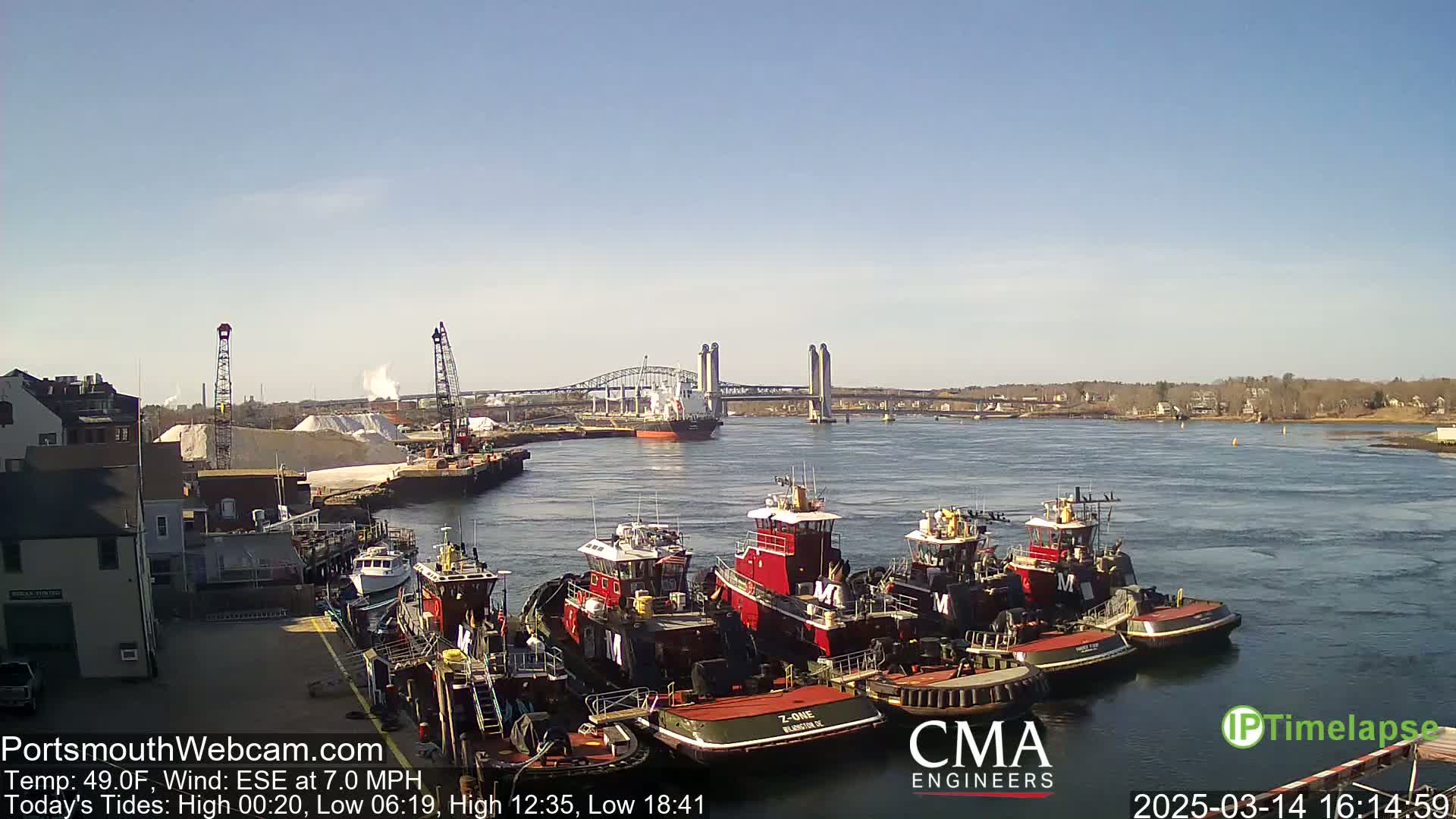 A sunny day shows several tugboats moored at a dock, with a larger vessel and a lift bridge in the background.