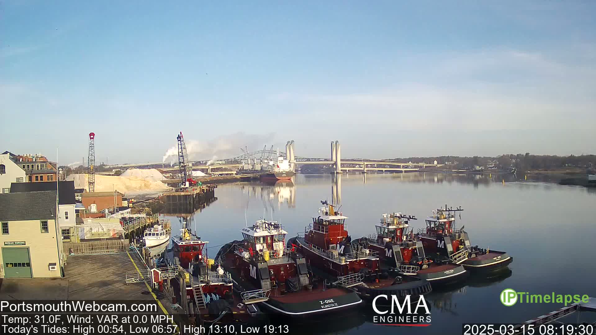Several tugboats are docked at a waterfront, with a large bridge and a cargo ship visible across a calm river under a clear sky.
