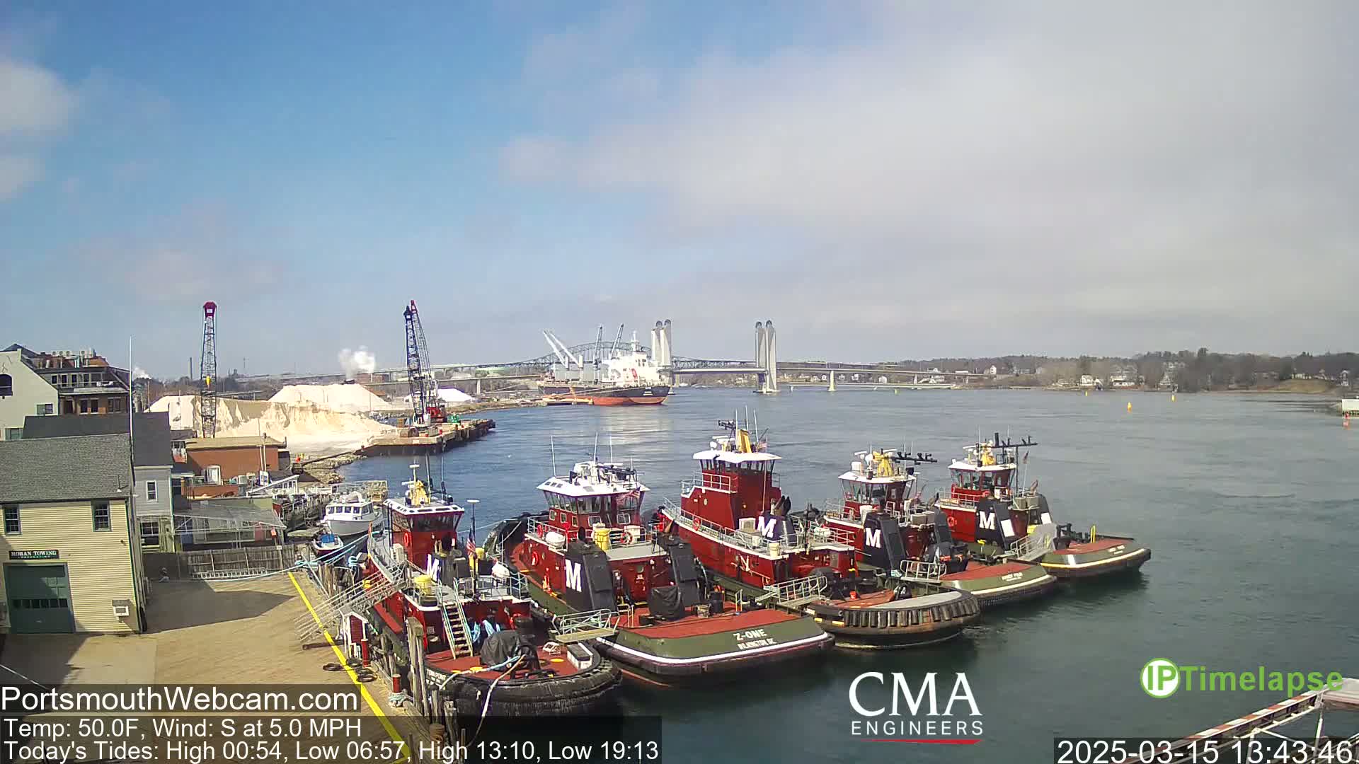 Several red tugboats are moored at a dock next to a large pile of white material, with a bridge and a cargo ship visible in the distance on a sunny day with a south wind.
