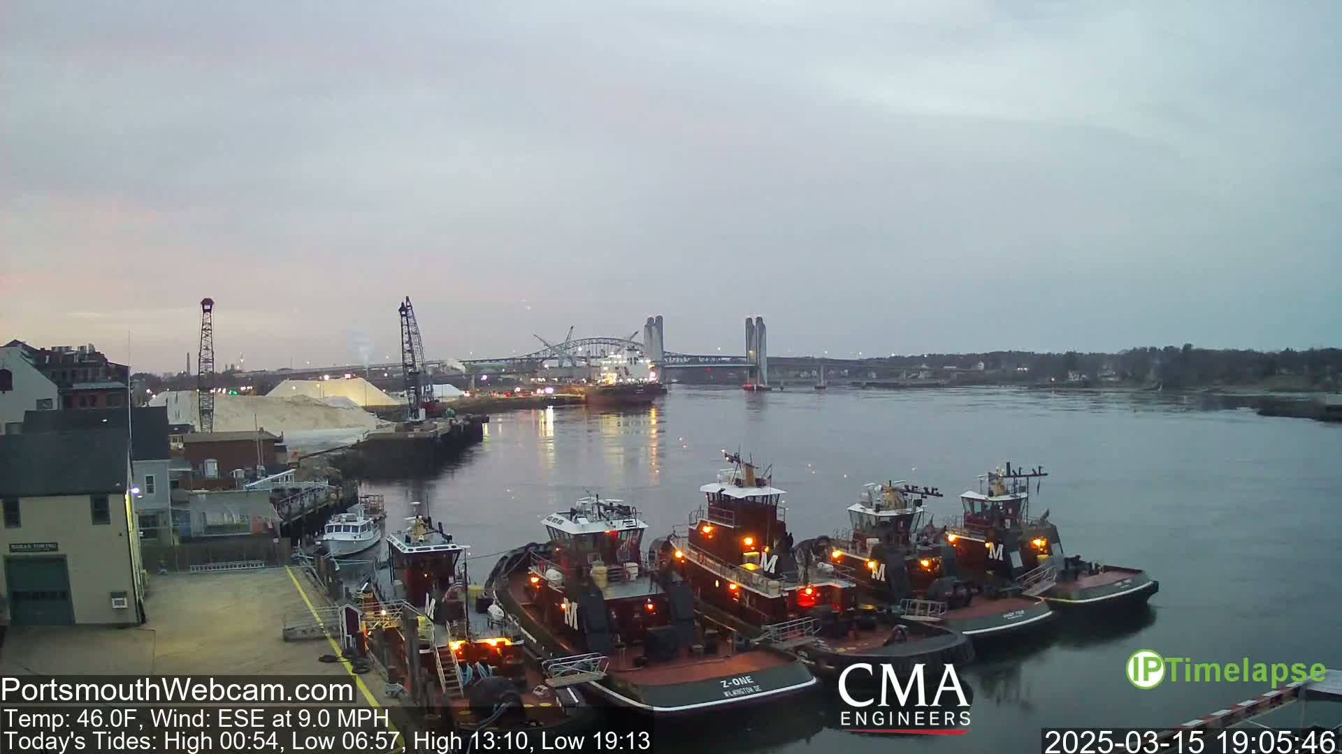 Several tugboats are moored at a dock next to a pile of white material, with a bridge and a large vessel visible in the distance under an overcast sky.