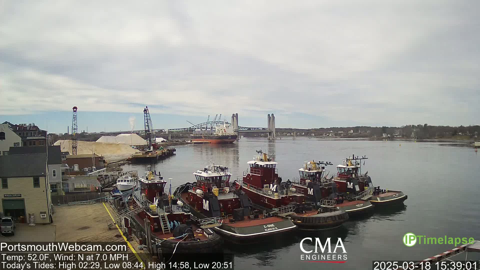 Several tugboats are moored at a dock next to a pile of sand, with a large cargo ship and a bridge in the background under a cloudy sky.