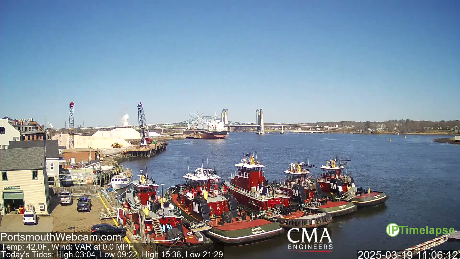 A clear, sunny day shows several tugboats docked near a large cargo ship under a bridge, with a pile of white material on the shore.