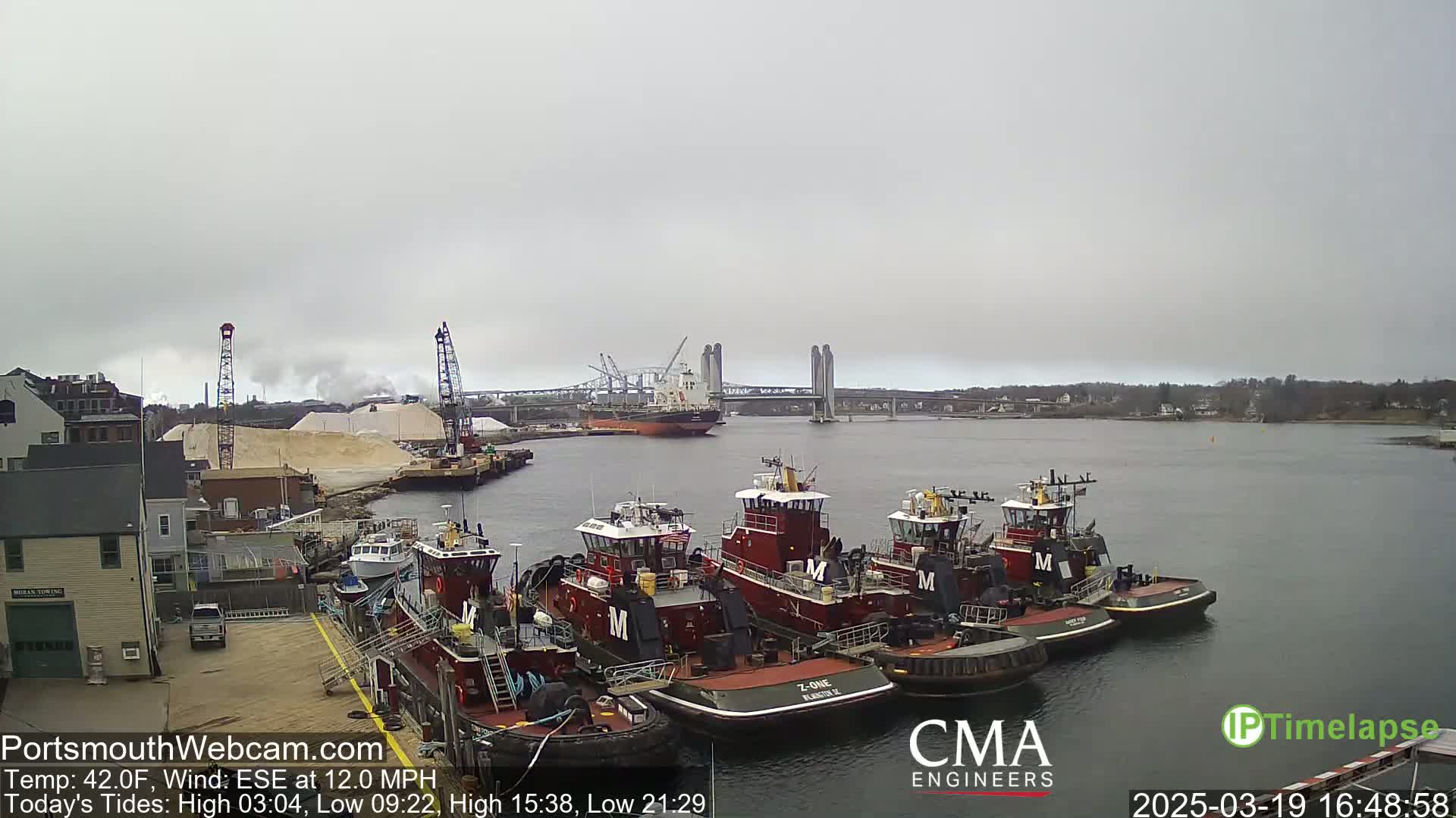 Several tugboats are moored at a dock next to a large pile of sand, with a cargo ship and a bridge visible in the distance under an overcast sky.