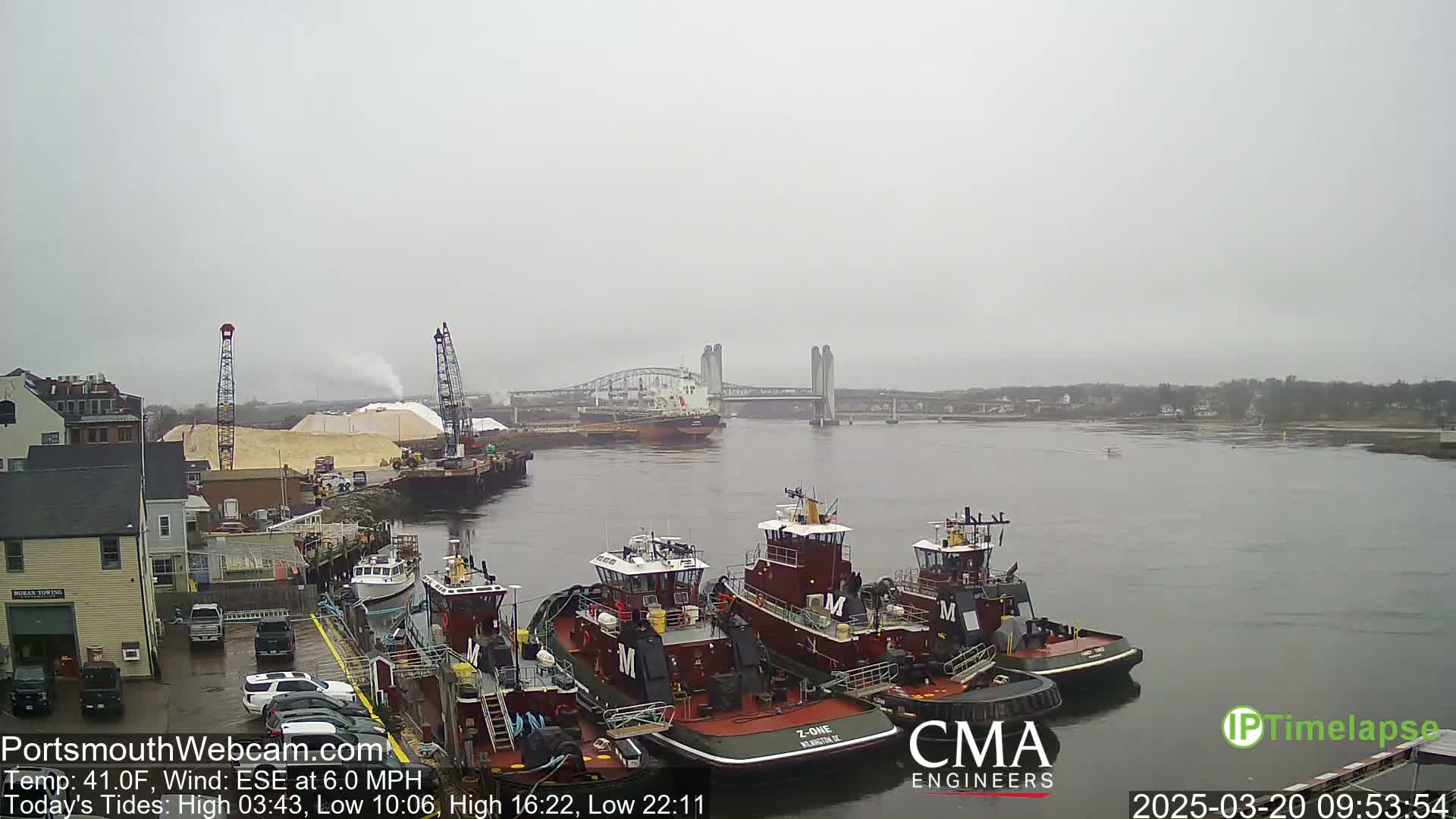 Several tugboats are docked at a waterfront location on an overcast day, with a large cargo ship and a bridge visible in the distance.