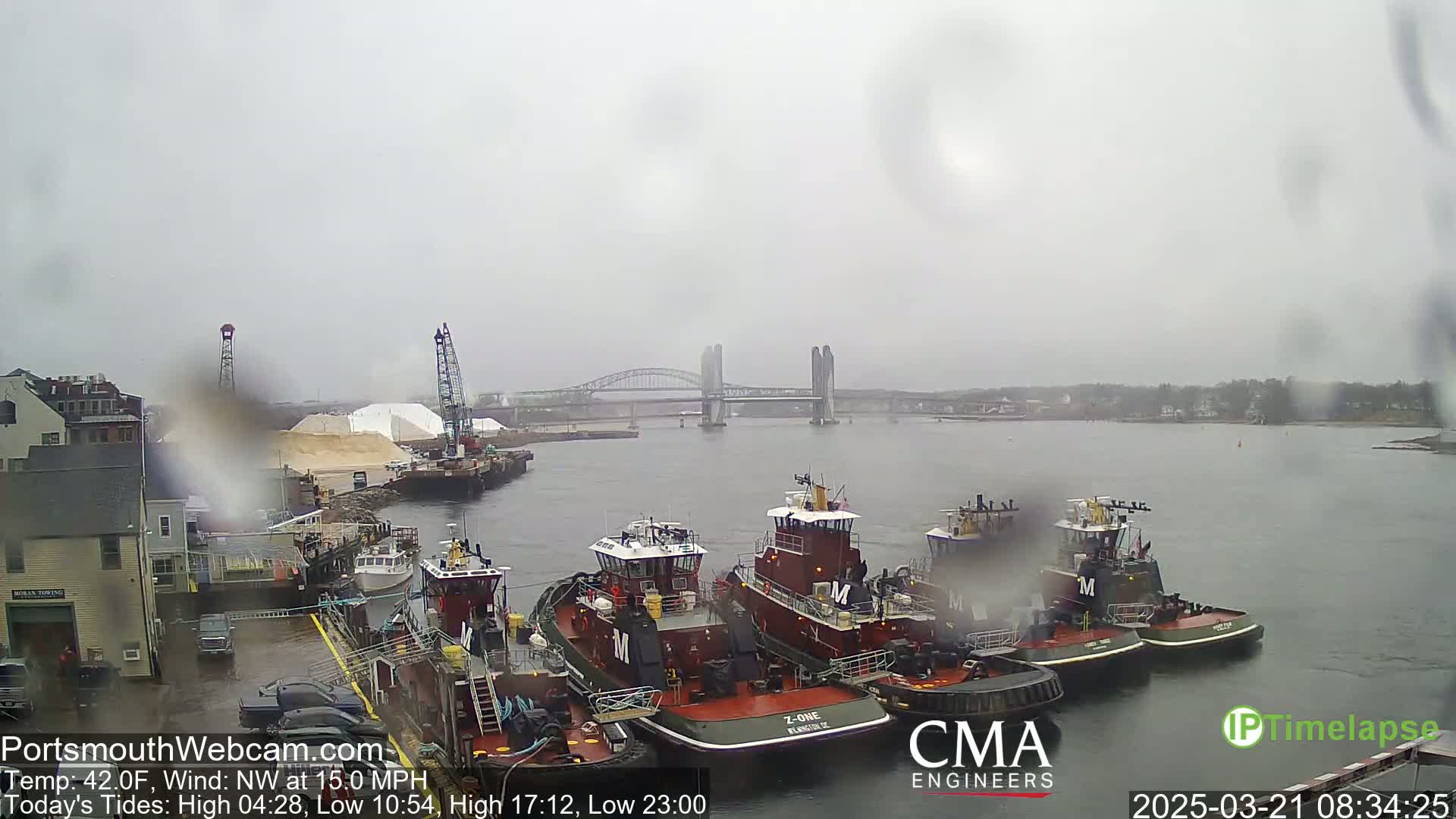 Several tugboats are moored at a dock in rainy weather, with a bridge visible in the background.