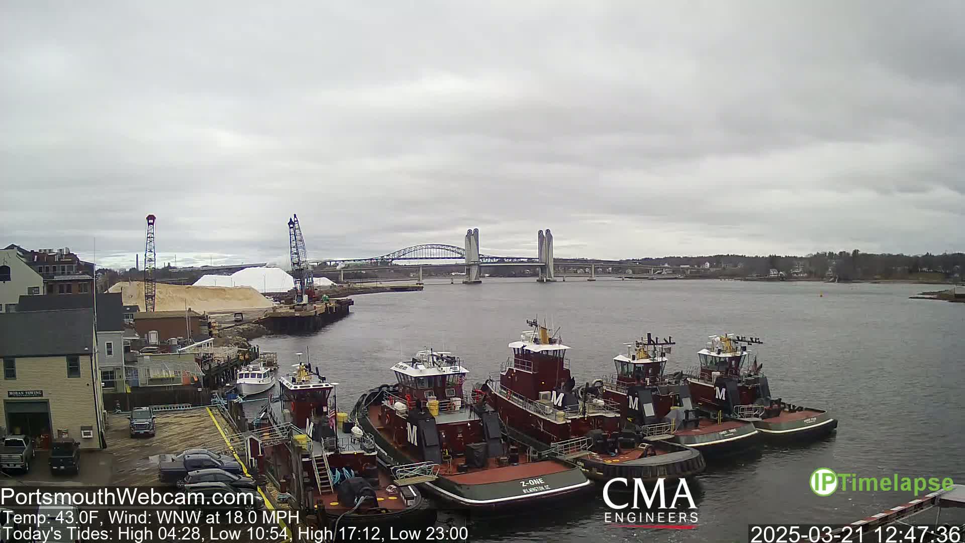Several tugboats are moored at a dock next to a large pile of sand or salt, with a bridge in the background under an overcast sky.
