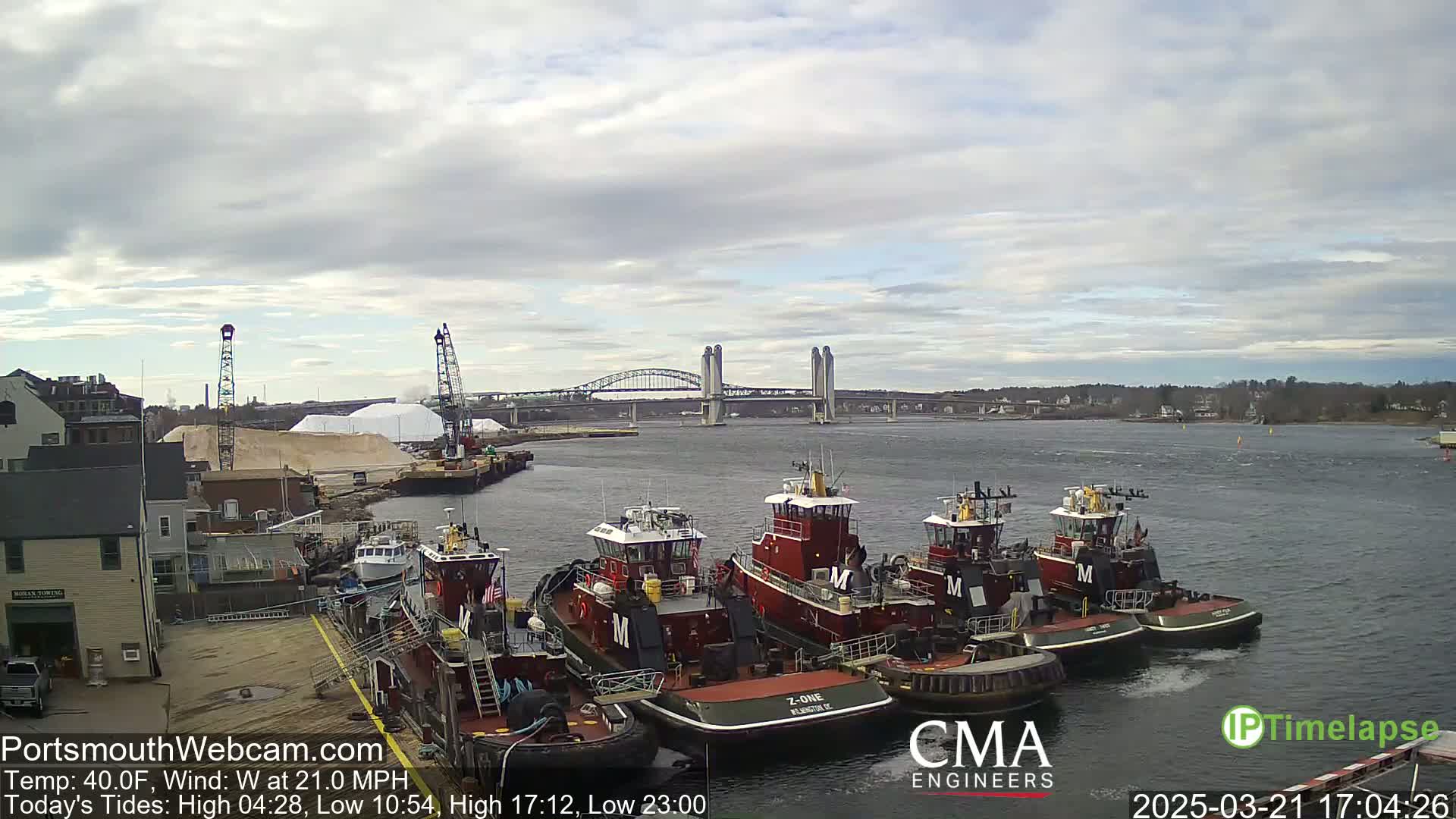 Several tugboats are moored at a dock near a bridge under a cloudy sky.