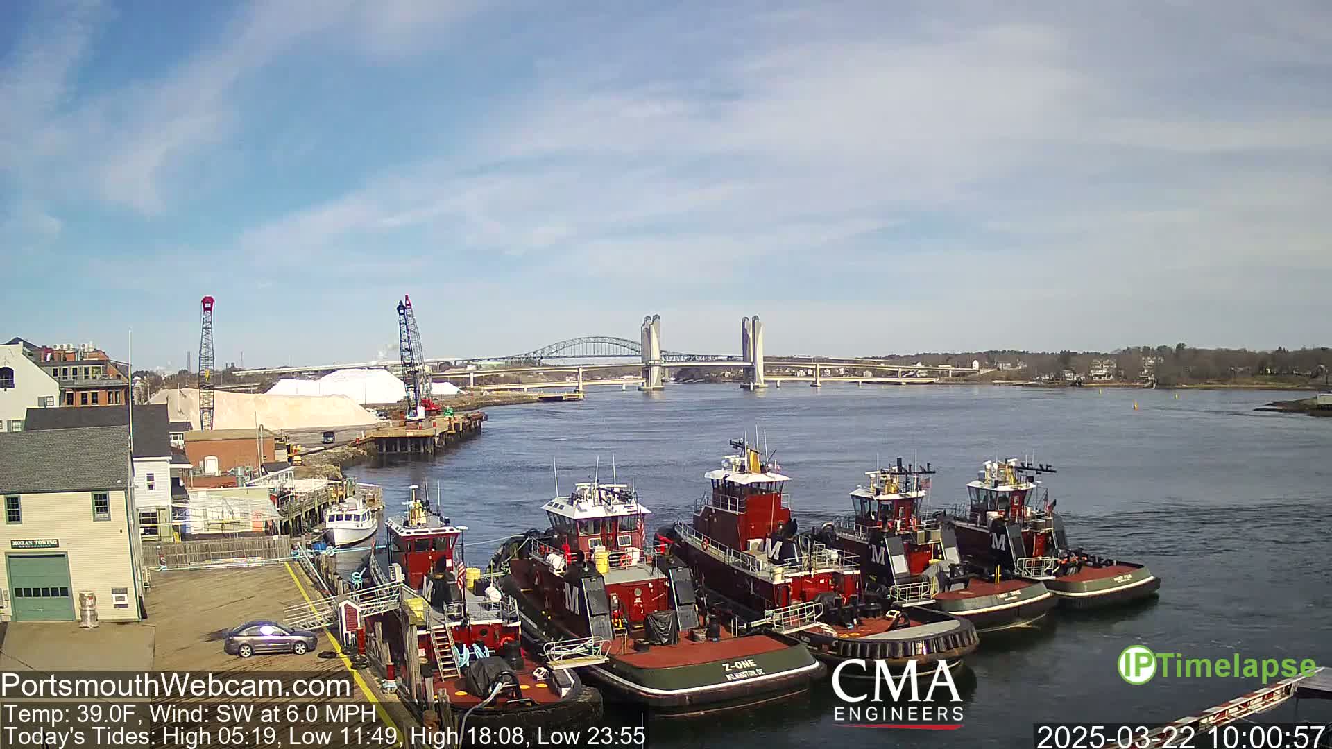 Under a partly sunny sky, several tugboats are docked at a waterfront with a large pile of white material nearby, and a double-leaf bascule bridge in the distance.
