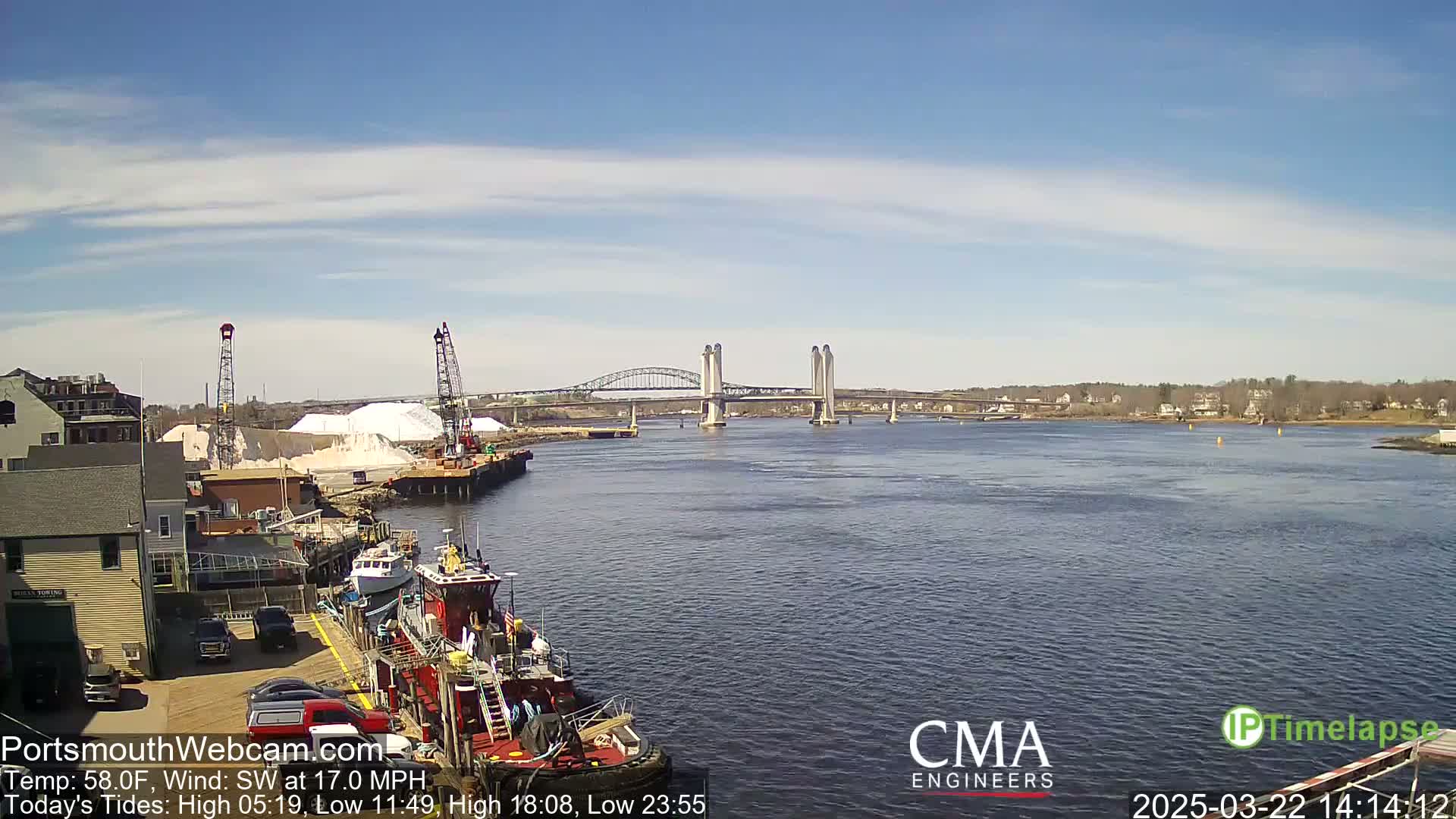 A tugboat is moored at a dock near a bridge spanning a river under a partly cloudy sky.