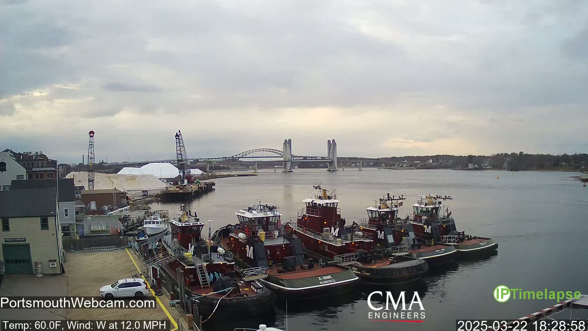 Several tugboats are docked at a waterfront, with a large pile of salt and a bridge in the background, under a cloudy sky.