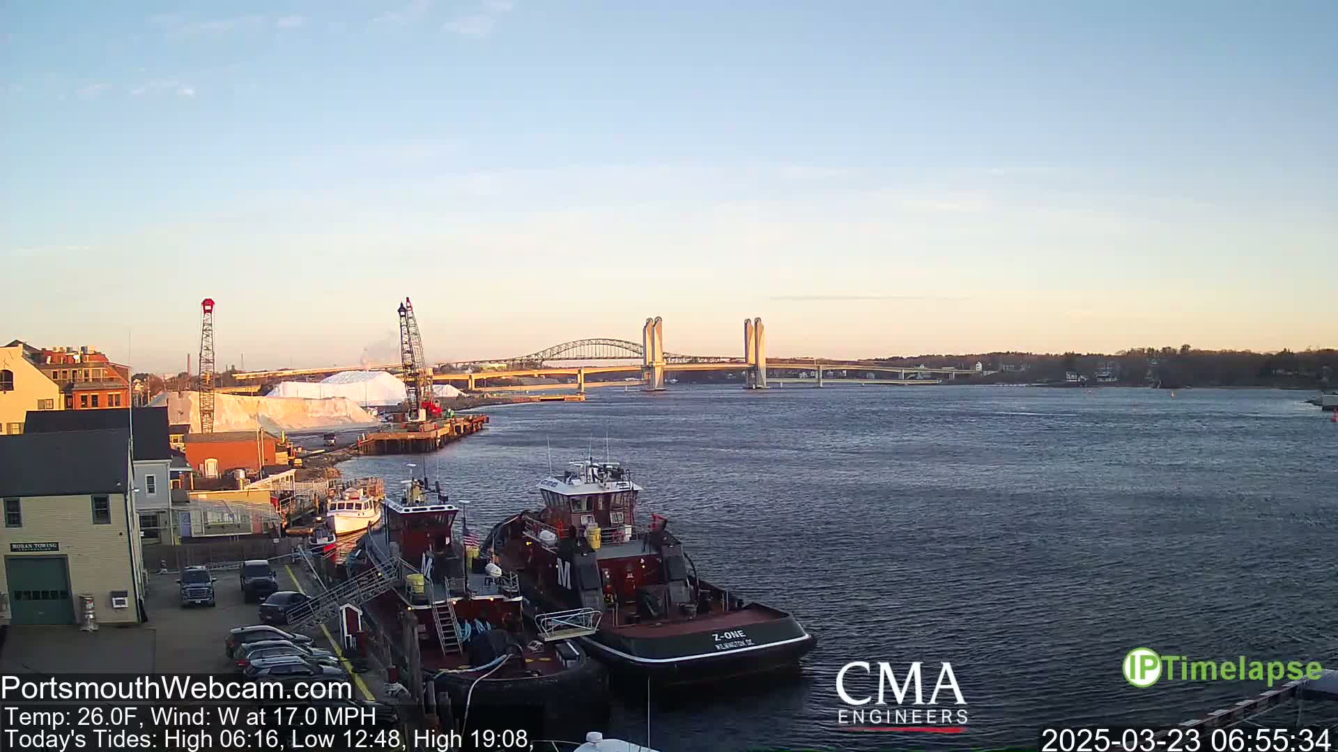 A harbor scene at sunrise, with two tugboats docked near a waterfront building and a large pile of white material, all under a clear sky and a light westerly wind.