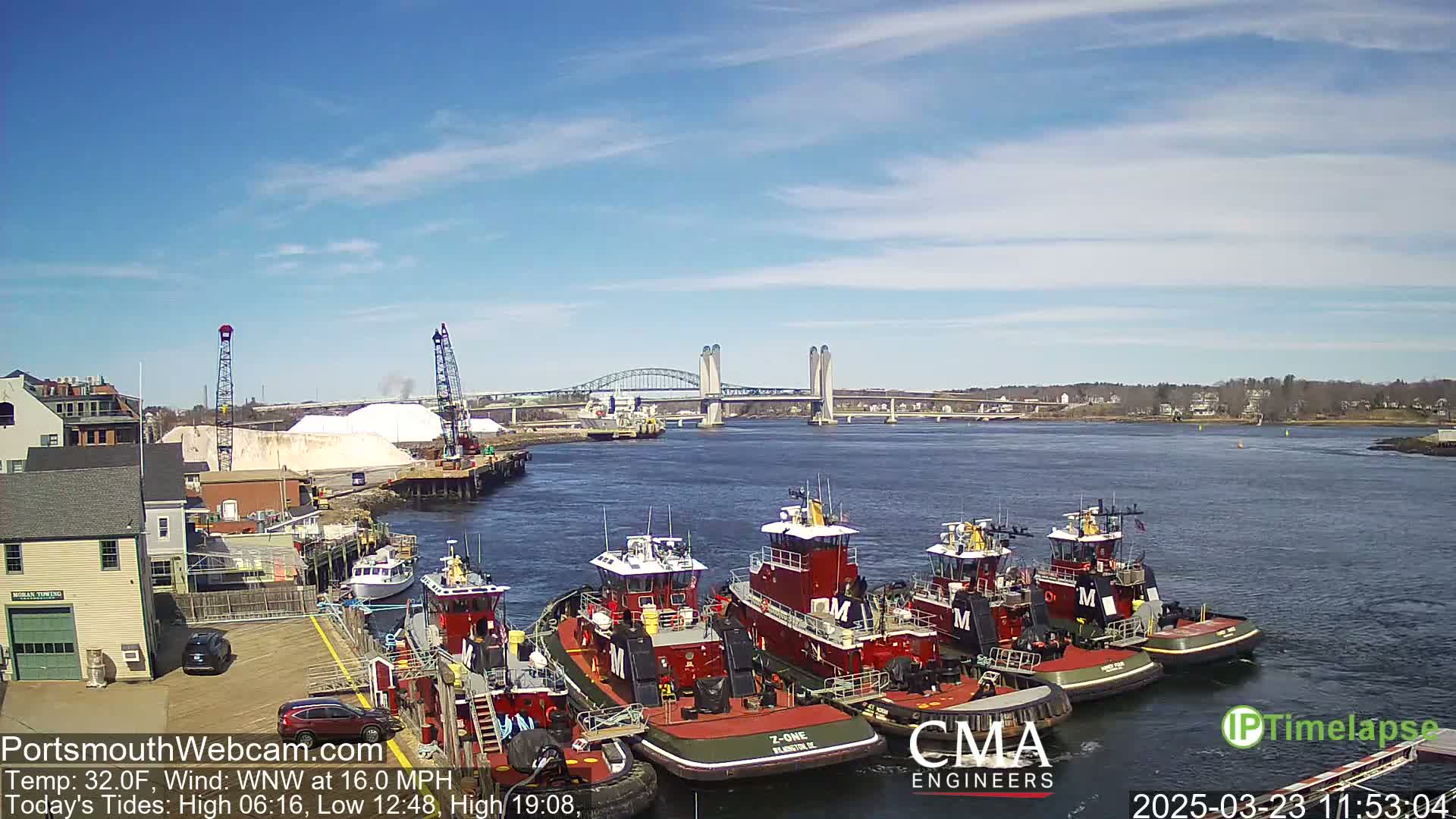 Several tugboats are moored at a dock next to a body of water under a mostly sunny sky, with a bridge and distant shoreline visible.