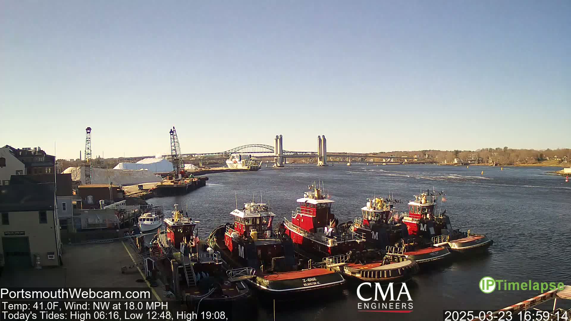 A harbor scene on a sunny day with a northwest wind at 18 mph shows several tugboats docked near a large pile of white material, with a bridge spanning the waterway in the background.