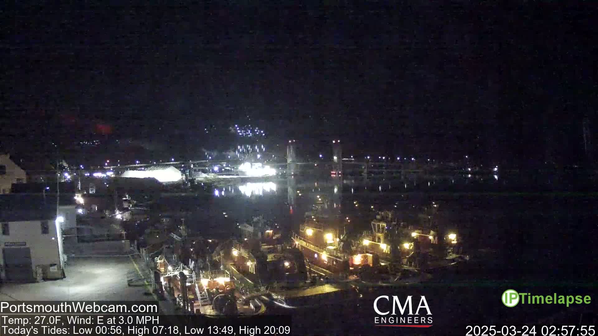 At night, several tugboats are docked near a bridge, under a clear, dark sky with an east wind.