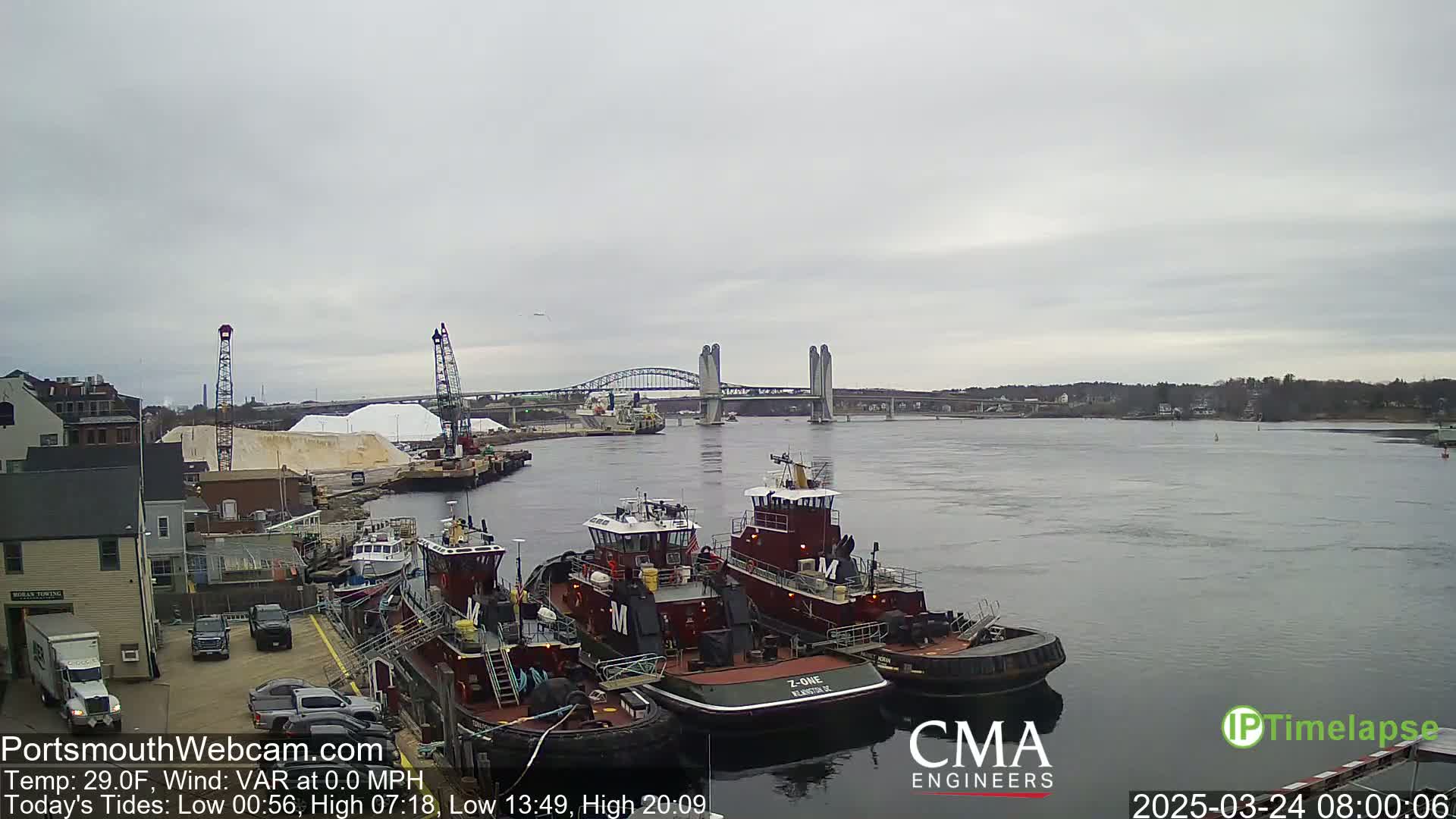Several tugboats are moored at a dock next to a large pile of salt, with a bridge spanning a river in the background under an overcast sky.