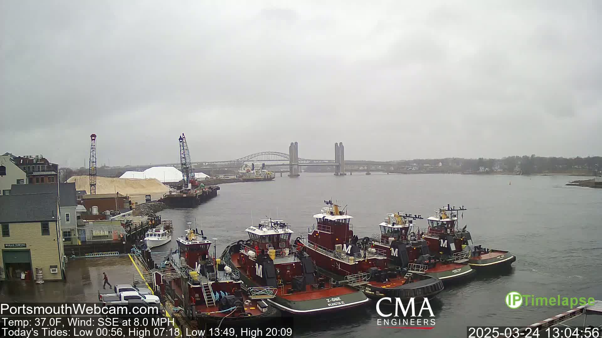 Several tugboats are moored at a dock next to a pile of sand, with a bridge visible in the distance under an overcast sky.