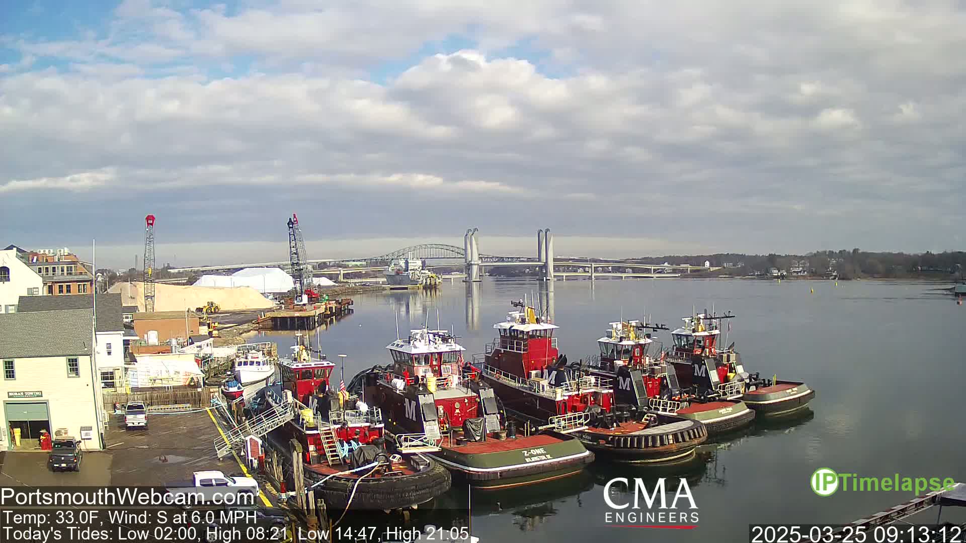 Several tugboats are docked at a wharf near a bridge on a partly cloudy day.