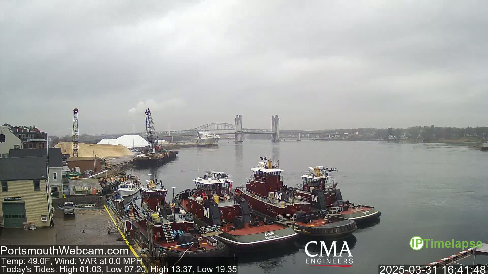 Several tugboats are docked at a waterfront, with a bridge and piles of sand visible in the distance under an overcast sky.