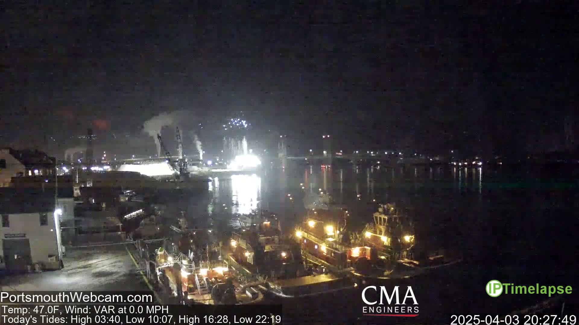 A nighttime view of a harbor with several tugboats, a brightly lit industrial area, and faint fireworks in the distance under a dark sky.