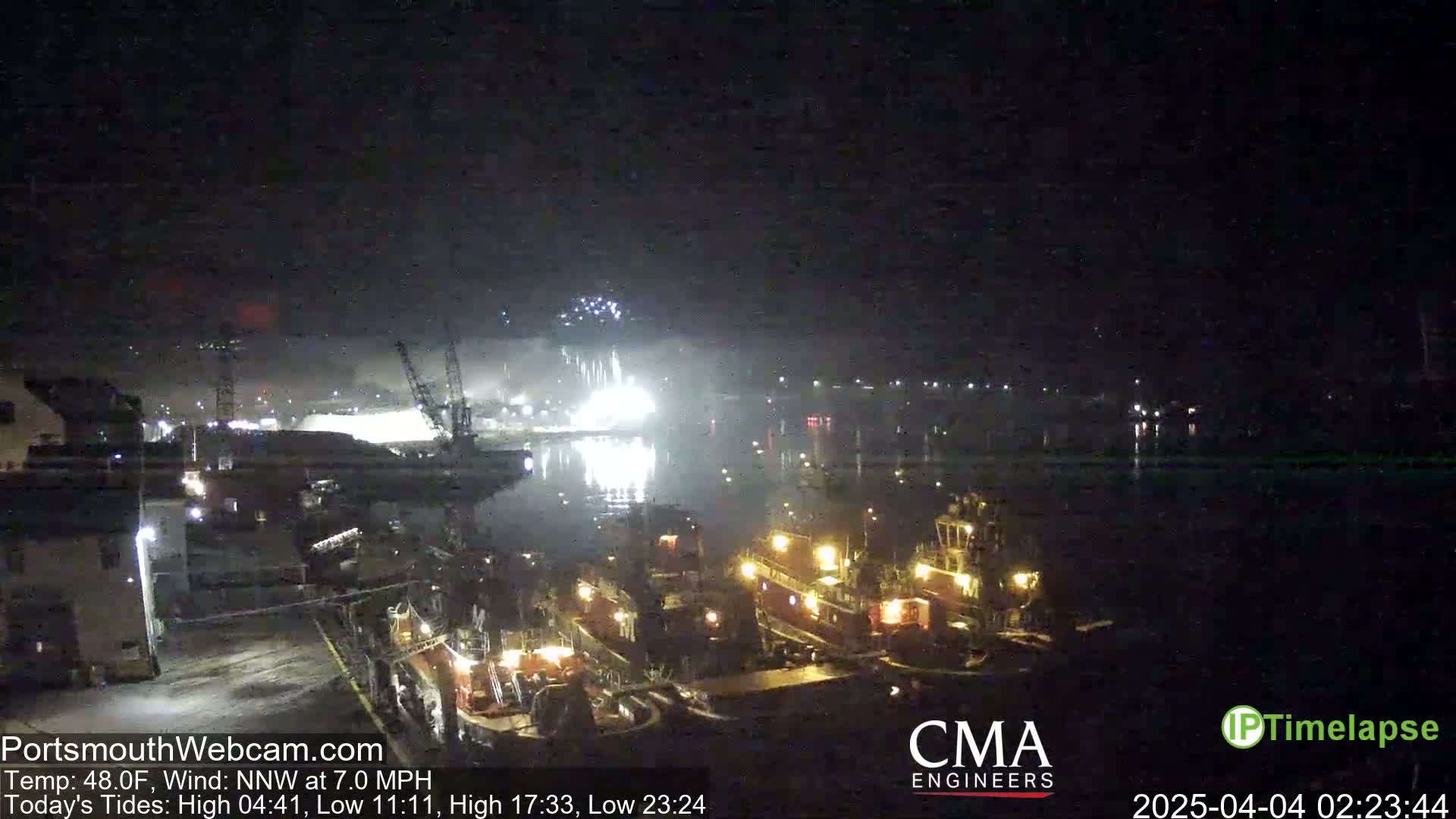 A nighttime harbor scene with fireworks exploding over the water, several tugboats are docked, and a light wind is blowing.