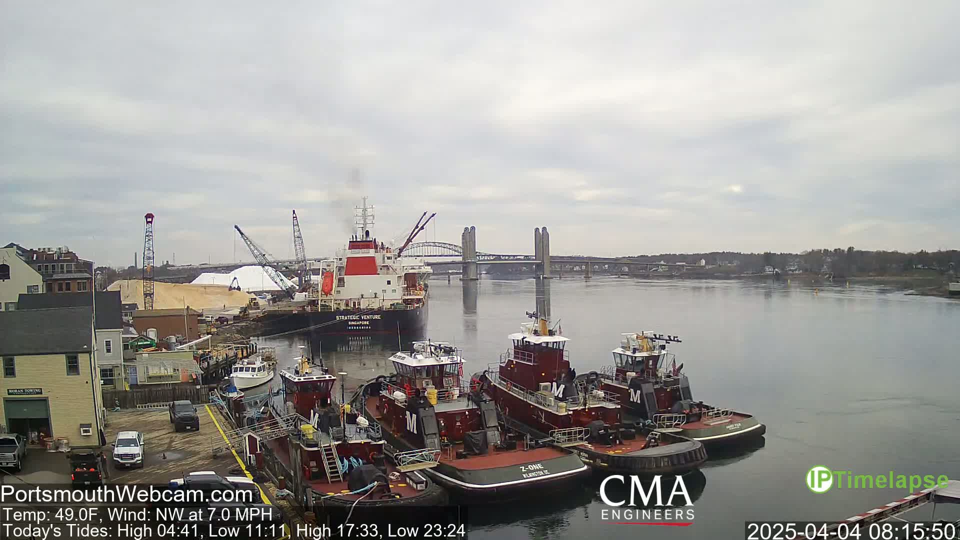 A large cargo ship is moored near several tugboats in a harbor under a cloudy sky, with a bridge visible in the distance.