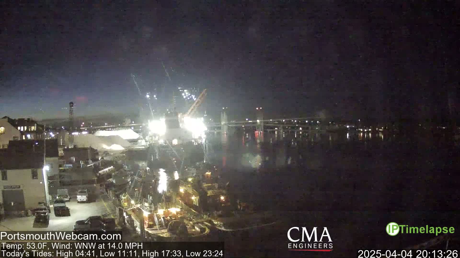 A nighttime view of a harbor with a bridge in the background, illuminated by bright lights and sparks, under a dark sky.