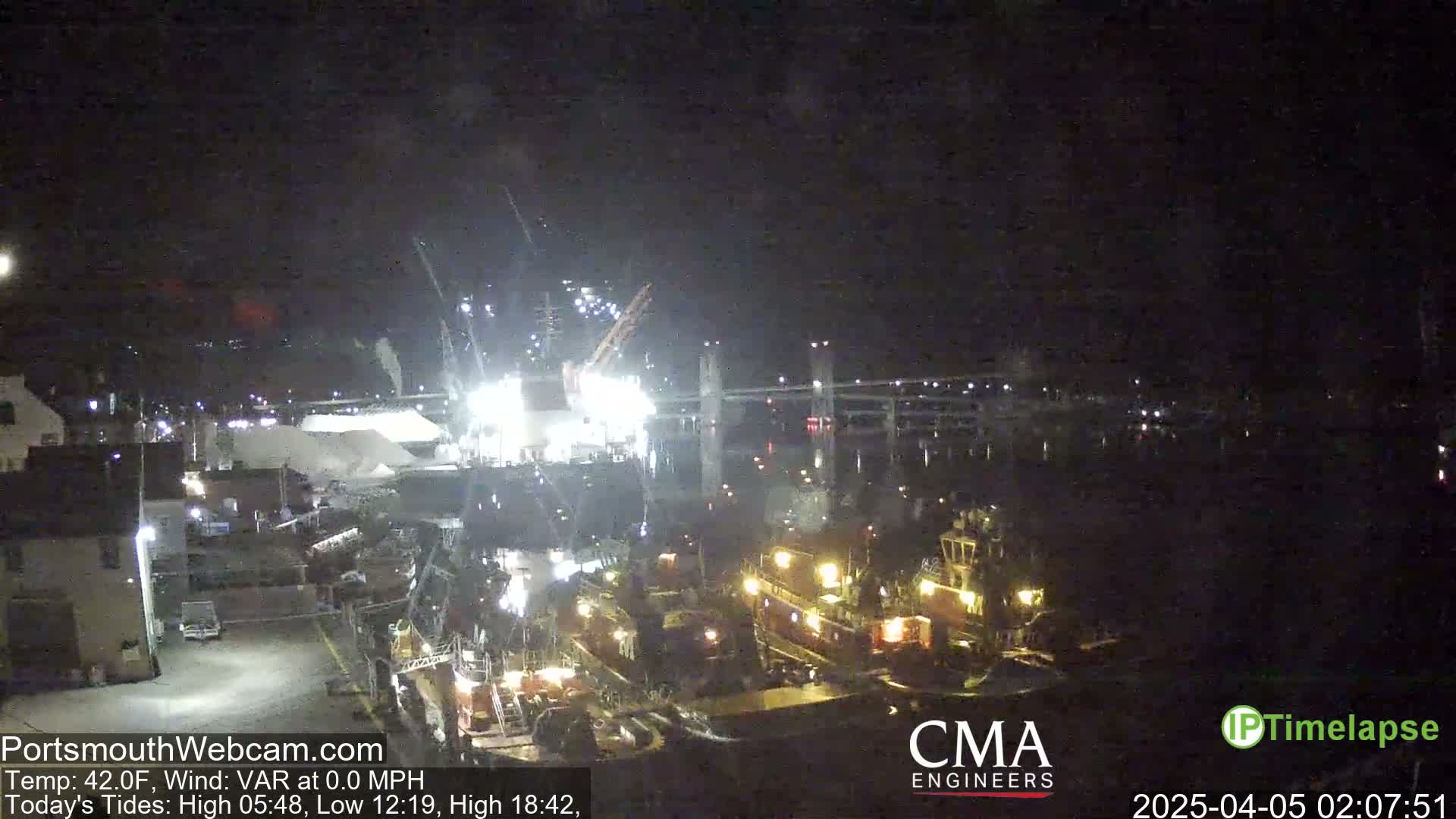 At night, brightly lit tugboats are moored near a large pile of material at a dock, with a bridge visible in the background under a dark, clear sky.