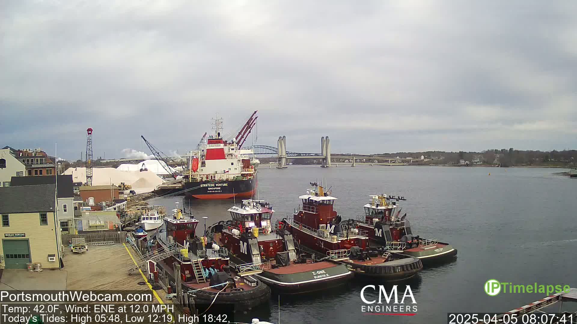Several tugboats are moored at a dock next to a large cargo ship and a bridge, under a cloudy sky.